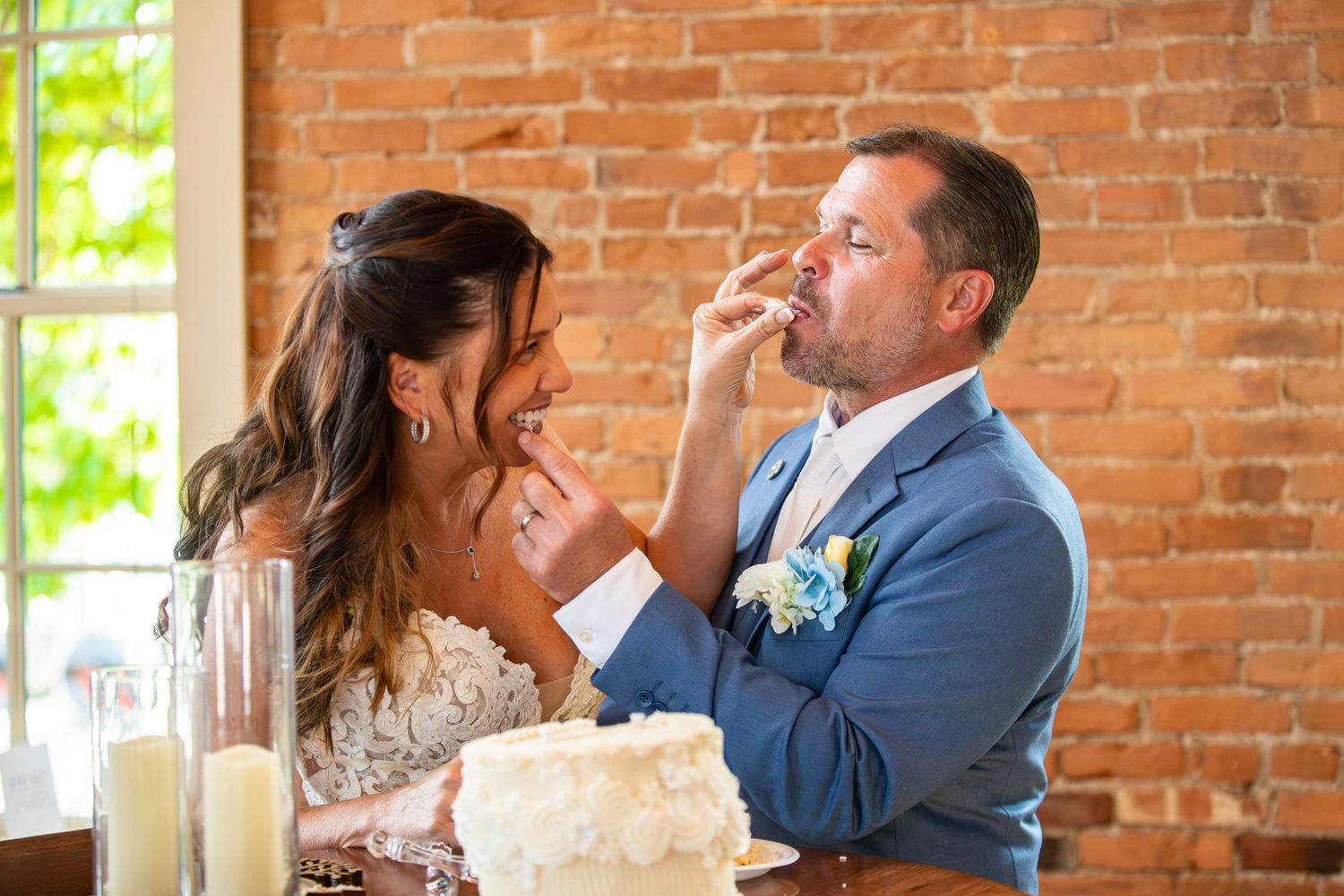 Bride feeding groom wedding cake at reception, brick wall background, white tiered cake on table.