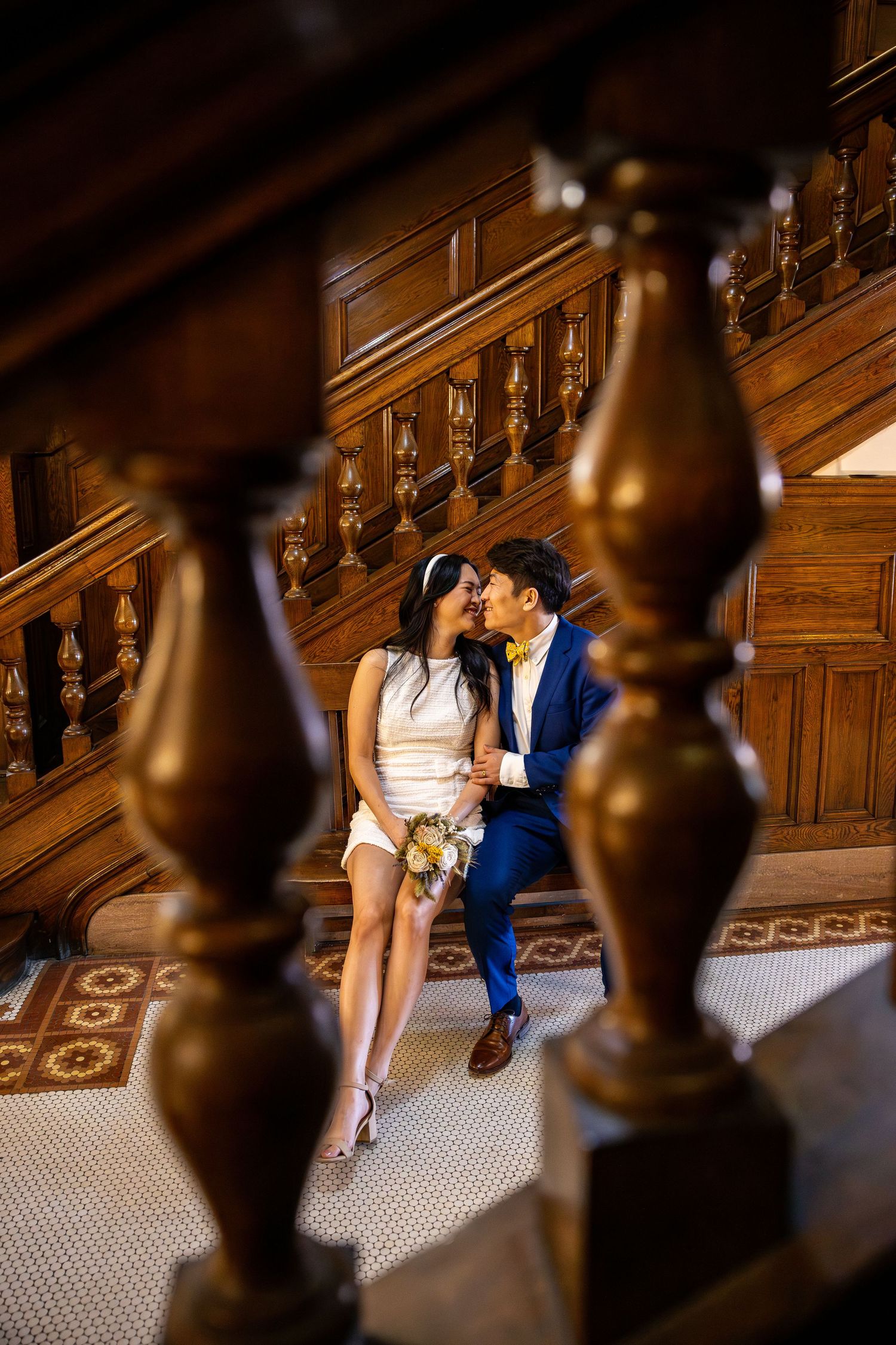 Bride and groom share a romantic moment seated on ornate wooden staircase in elegant venue.