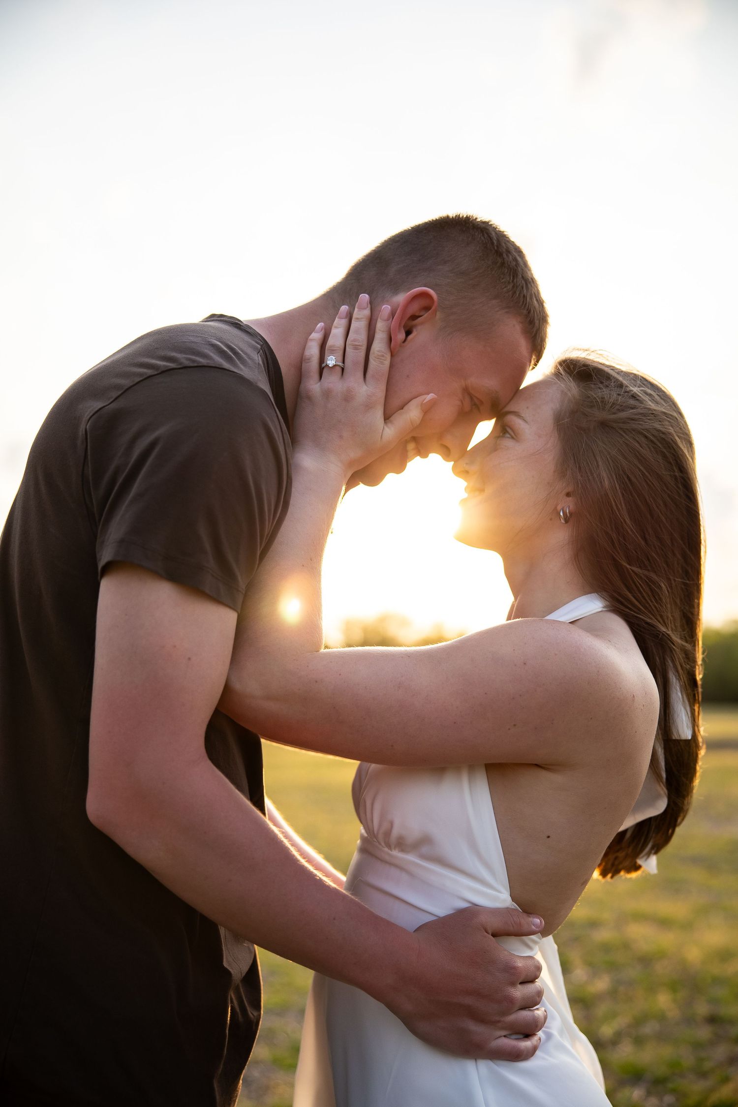 Couple sharing a romantic moment at golden hour sunset, woman in white dress with engagement ring visible.