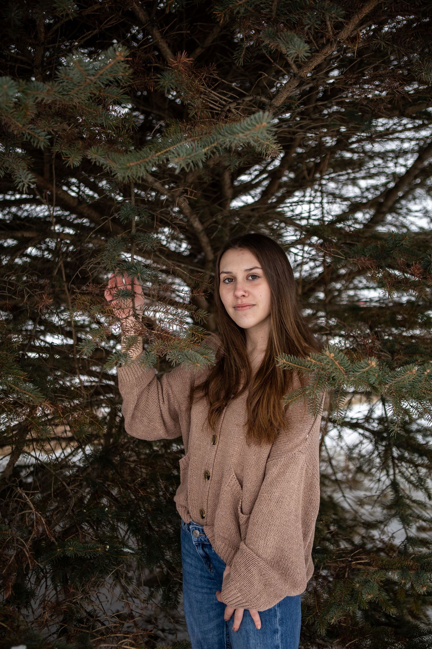 A woman in a beige sweater poses against evergreen trees in a winter outdoor photoshoot series.