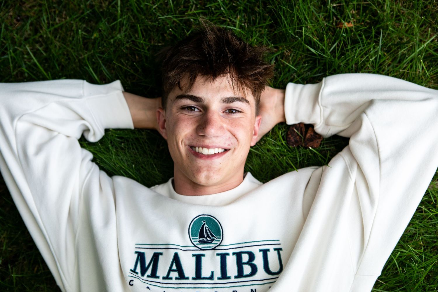 Someone wearing a white Malibu sweatshirt lies in green grass while smiling at the camera.