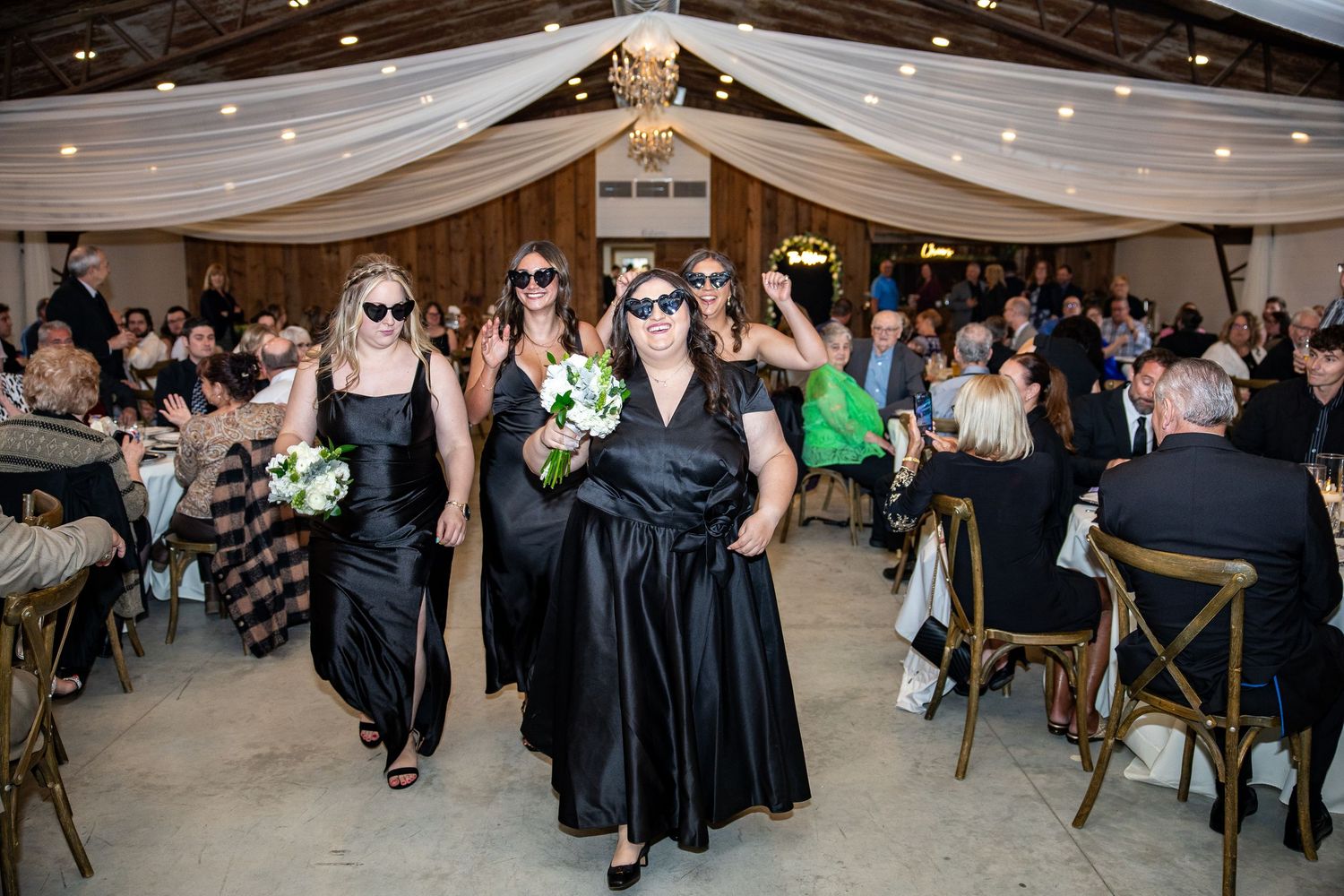 Bridesmaids in black gowns walk down wedding reception aisle holding white bouquets in elegant draped venue.