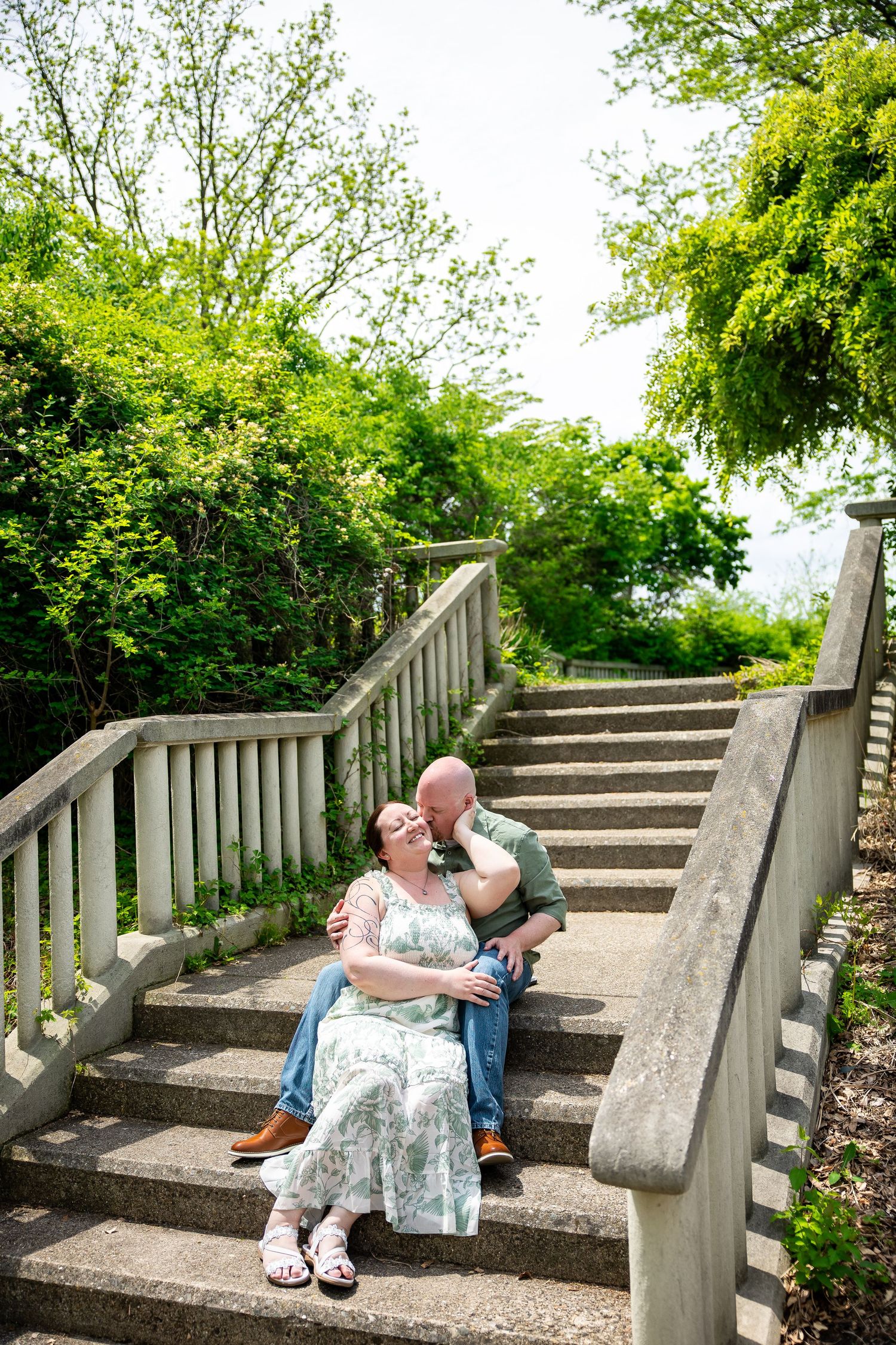 Couple sitting on stone steps surrounded by lush green trees, sharing a tender moment outdoors in spring.