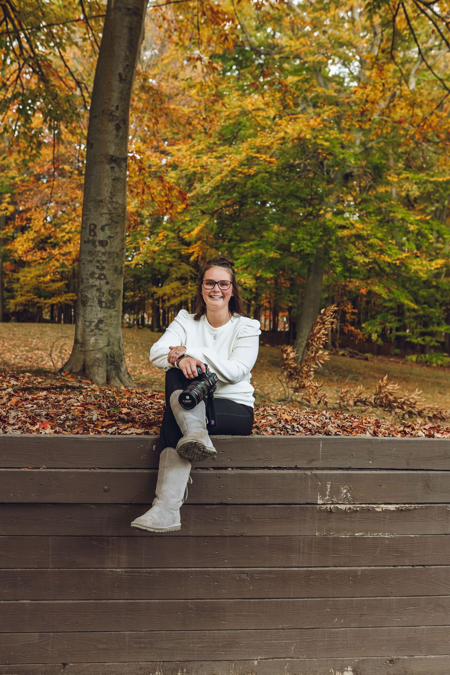 Smiling woman with glasses sits on a wooden wall holding a camera, surrounded by colorful autumn trees and fallen leaves.