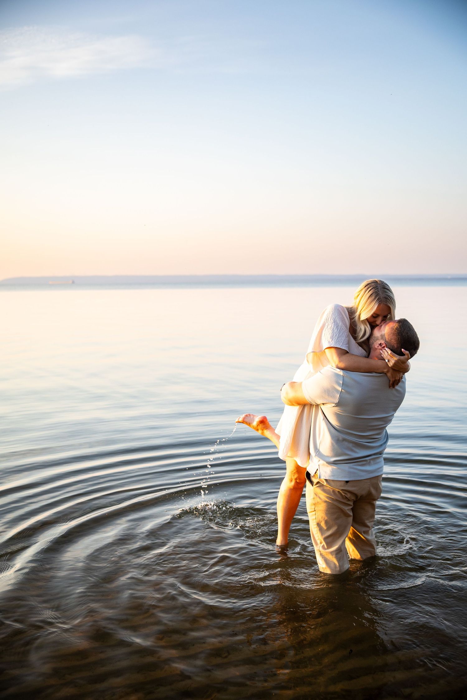 Couple embracing romantically while standing in shallow ocean water at sunset, with calm sea and pastel sky in background.