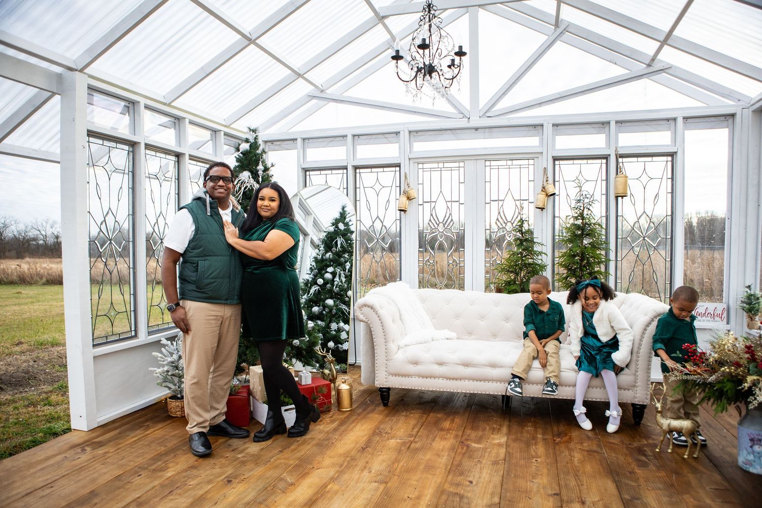 Family poses together in a bright sunlit conservatory decorated with Christmas trees and white furniture.