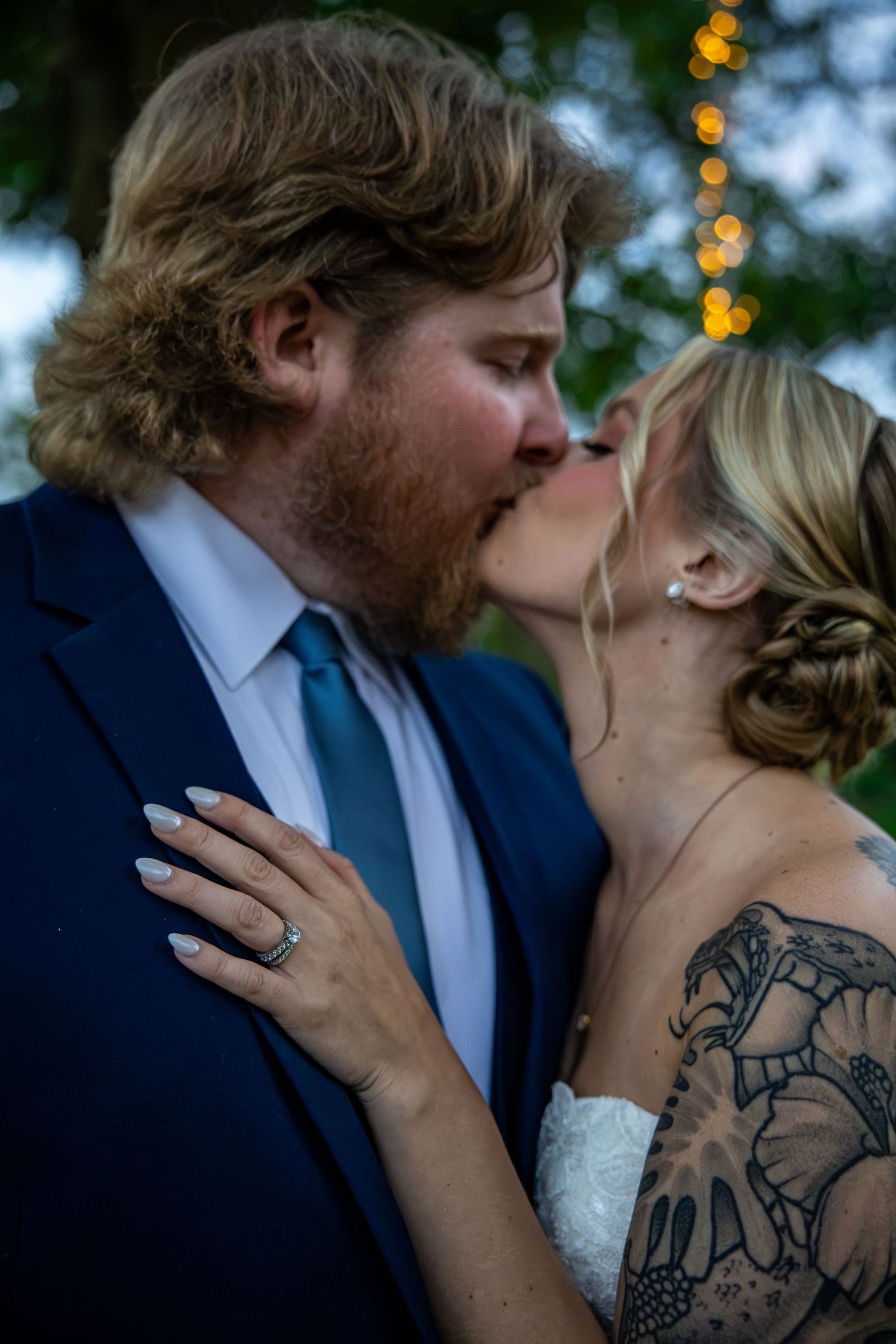 A bearded groom in a navy suit and teal tie kisses his bride, who wears a floral shoulder tattoo and a ring, with string lights behind.