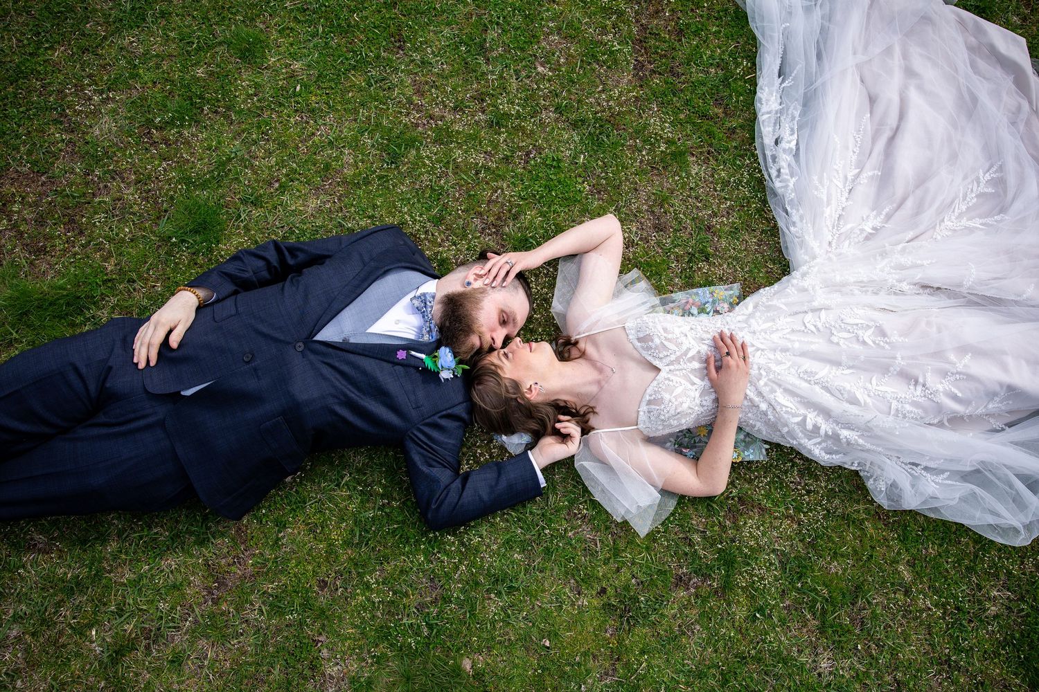 Bride and groom lying on green grass in wedding attire, sharing a romantic moment from aerial view.
