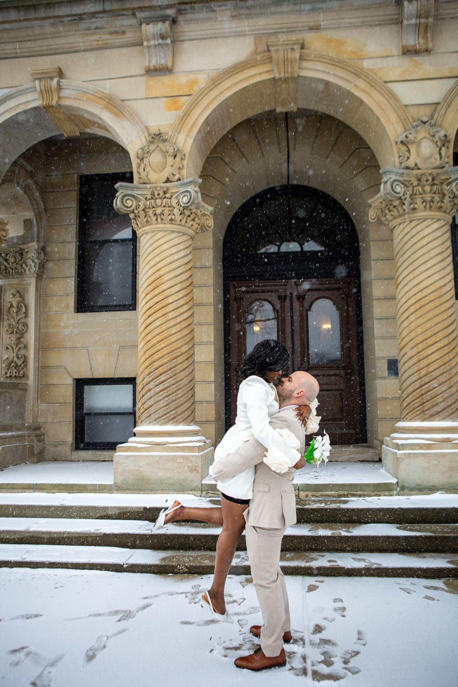 A couple shares a romantic embrace on snowy steps in front of an ornate stone building with twisted columns and arched doorway.