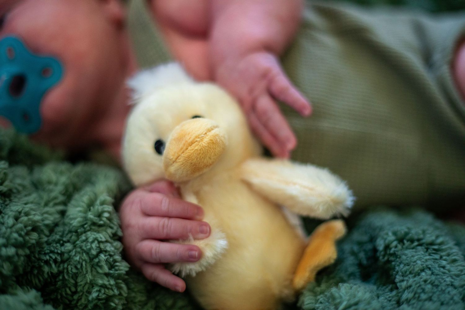 Baby hands reaching for yellow plush duck toy on green blanket.