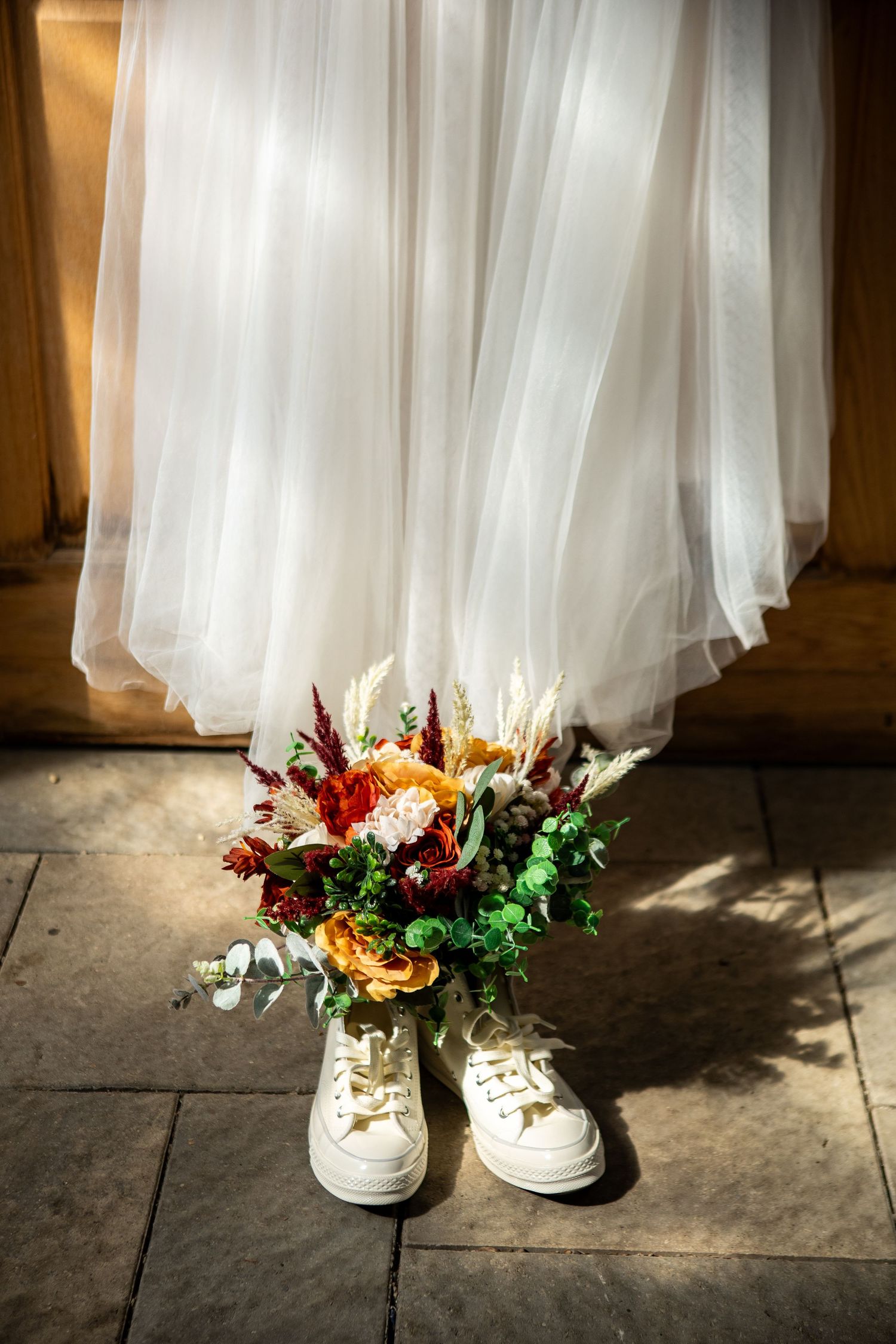 A pair of white sneakers filled with colorful wedding flowers sits beneath flowing tulle wedding dress fabric.