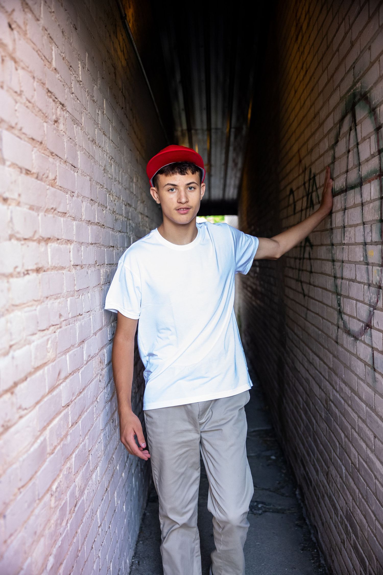 Young man in red cap and white t-shirt leans against brick wall in narrow urban alley with graffiti.