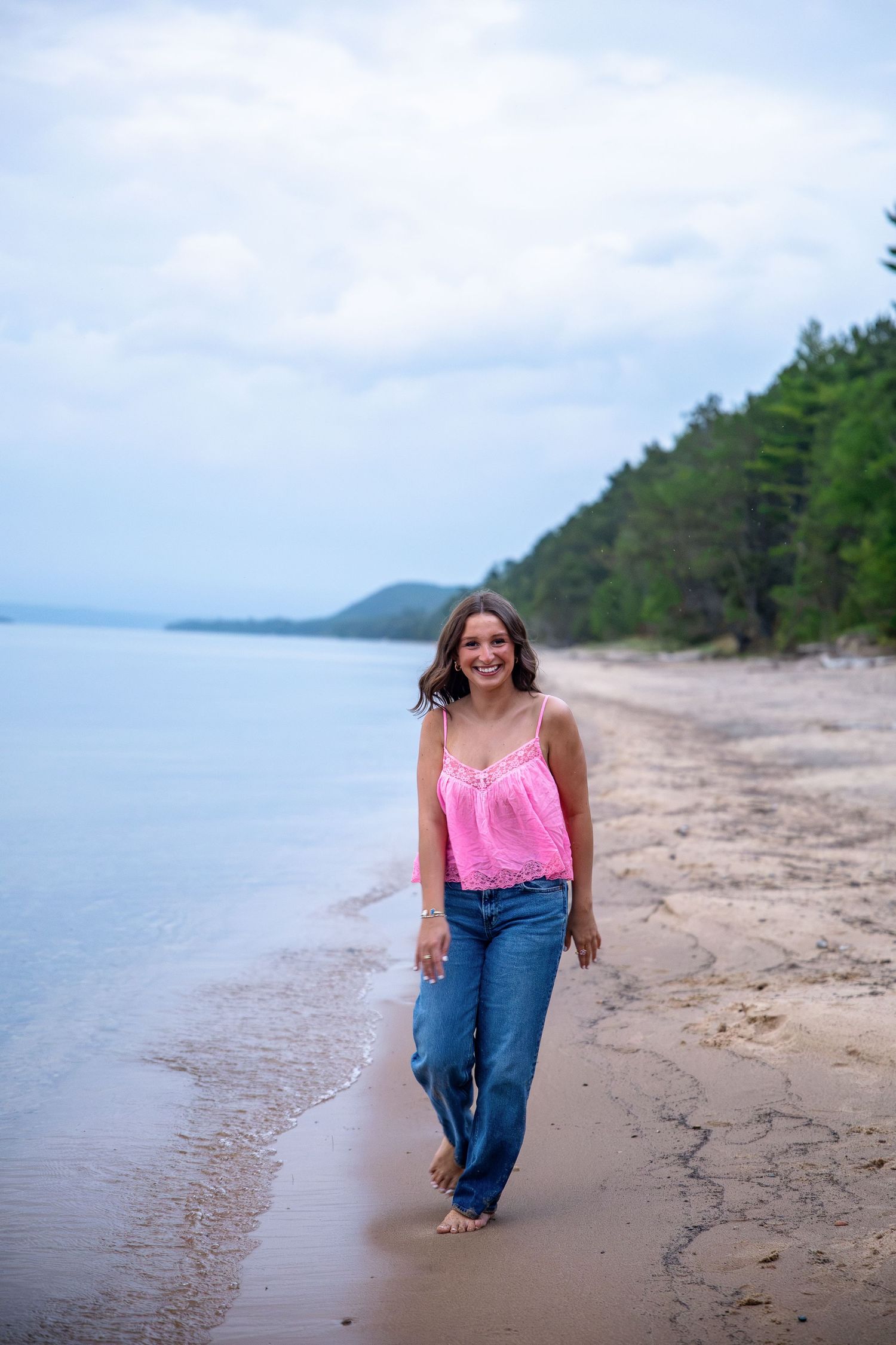 A smiling young woman in a pink tank top and jeans walks barefoot along a sandy beach with trees in the background.