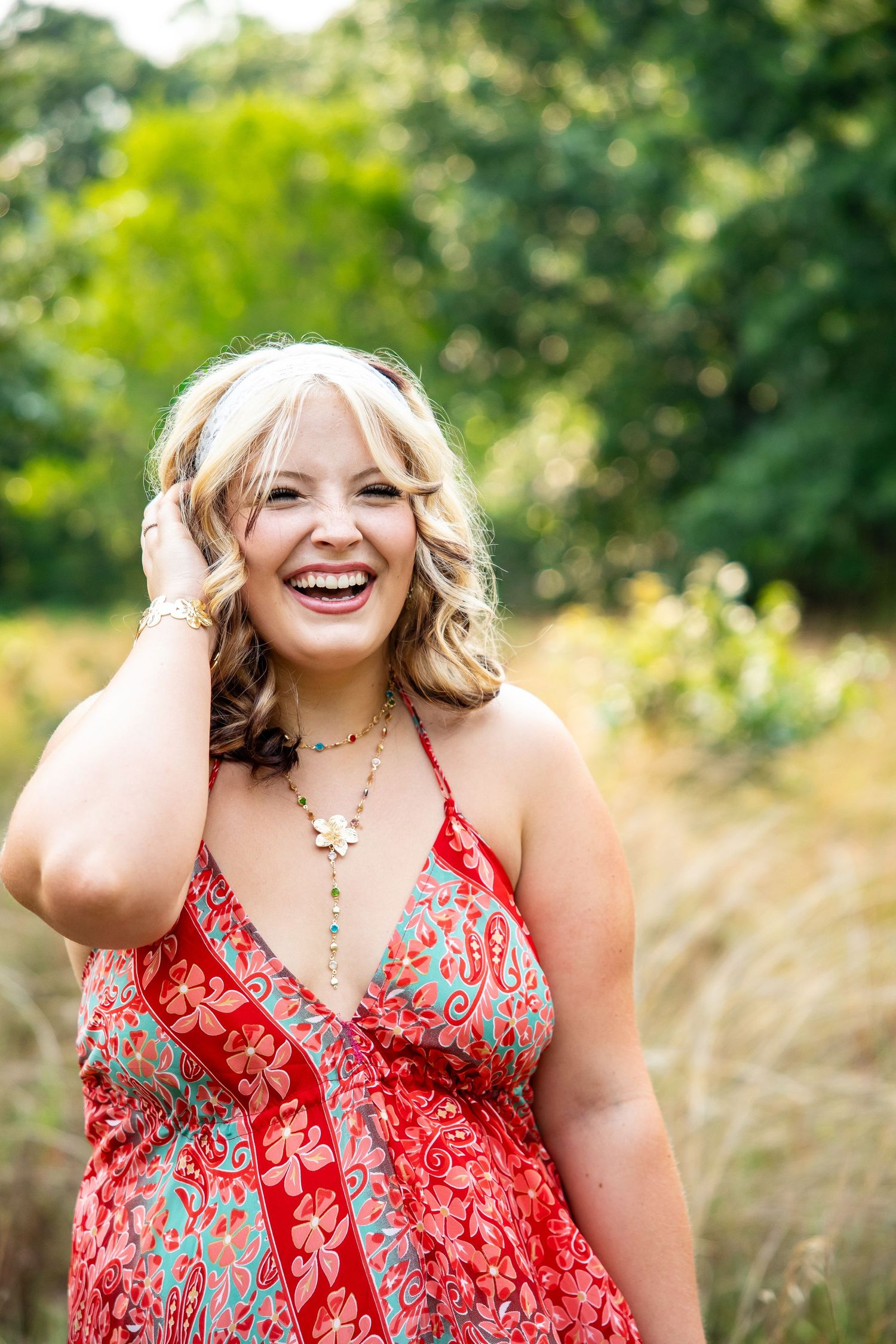 A smiling blonde woman in a red floral sundress laughs outdoors, hand in hair, wearing a layered necklace.