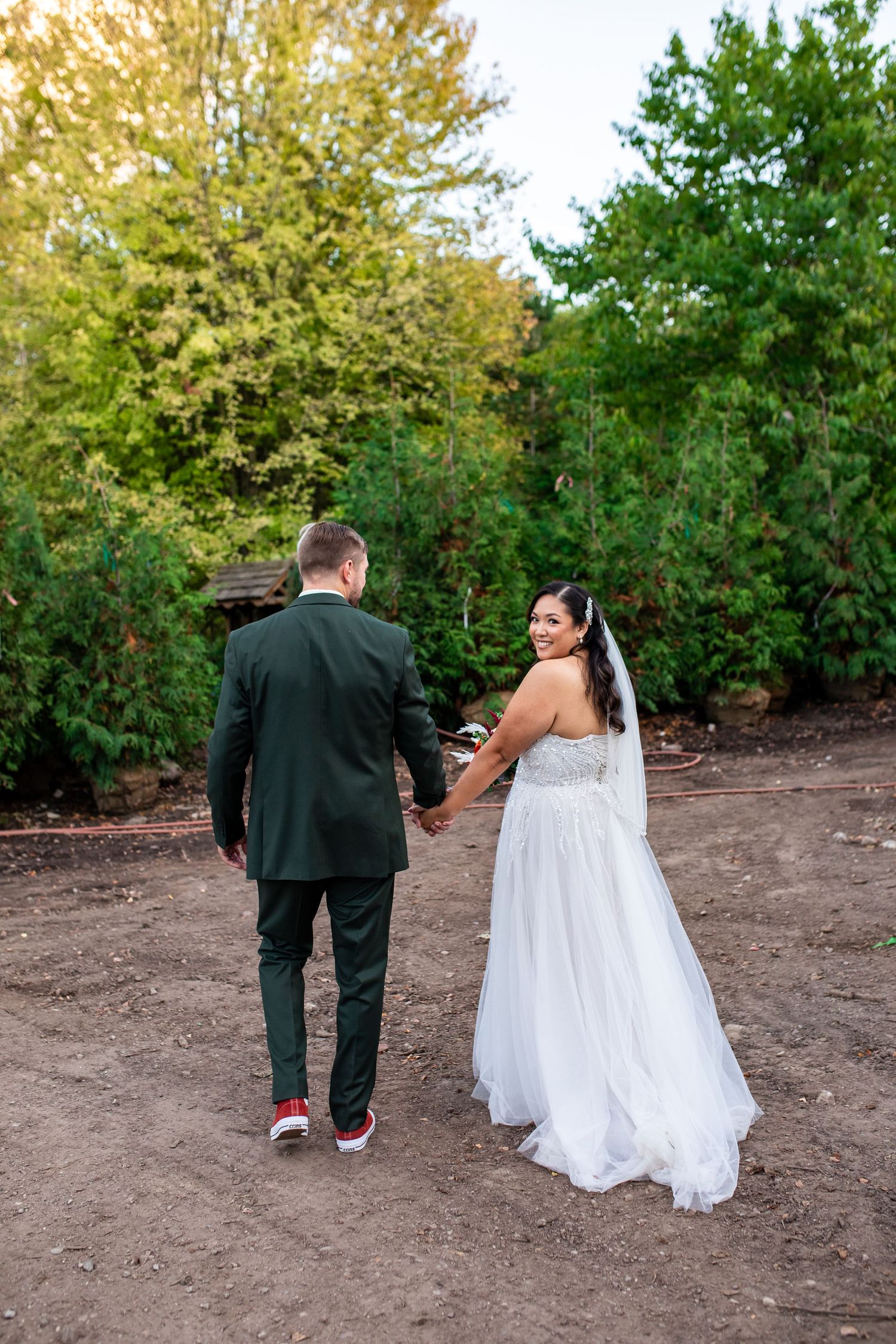 Bride in white gown and groom in dark suit walk hand-in-hand through a lush green garden on their wedding day.