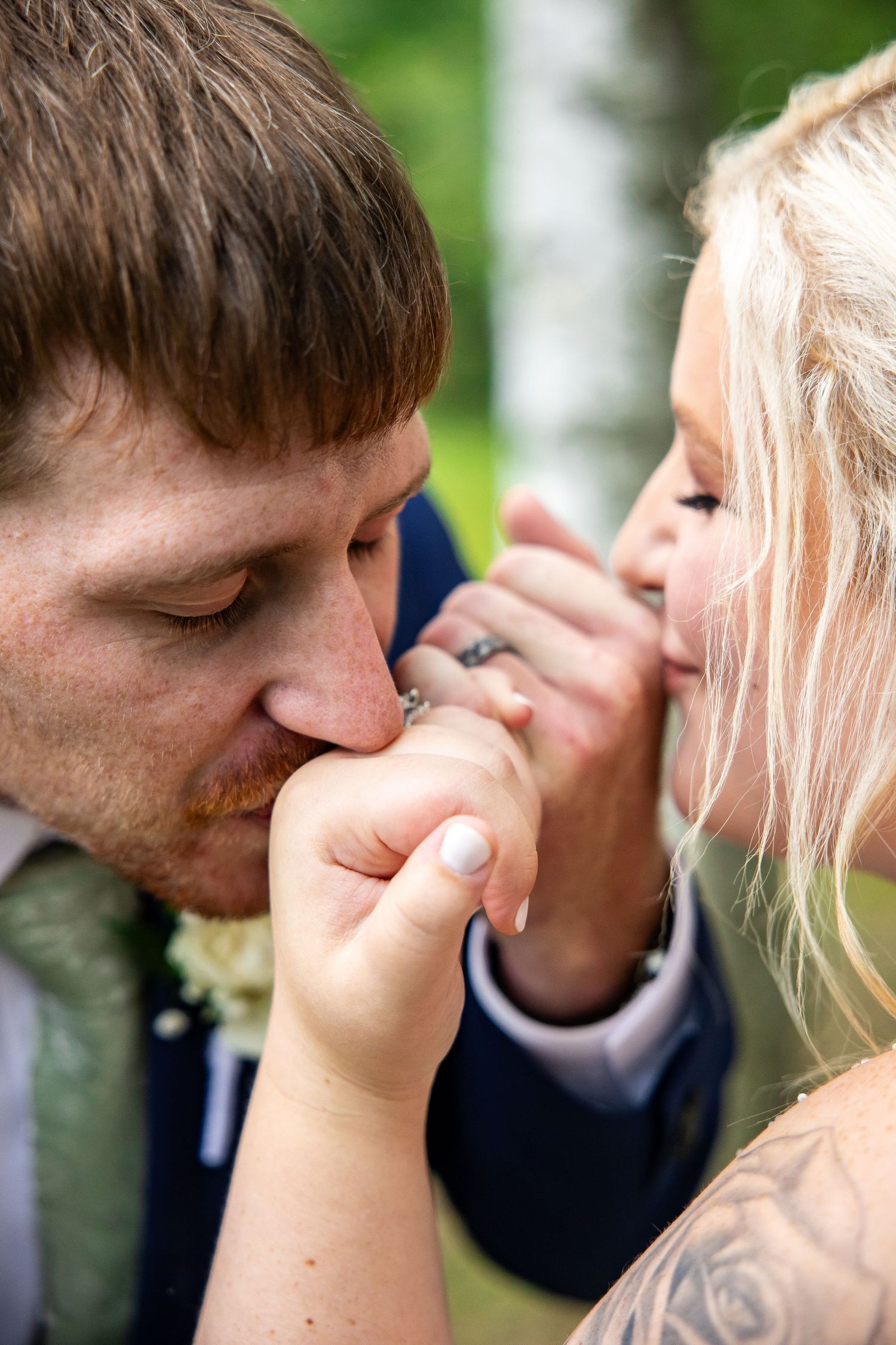 Groom in a navy suit and green tie tenderly kisses the hand of his blonde bride, showing her ring, in an outdoor wedding portrait.