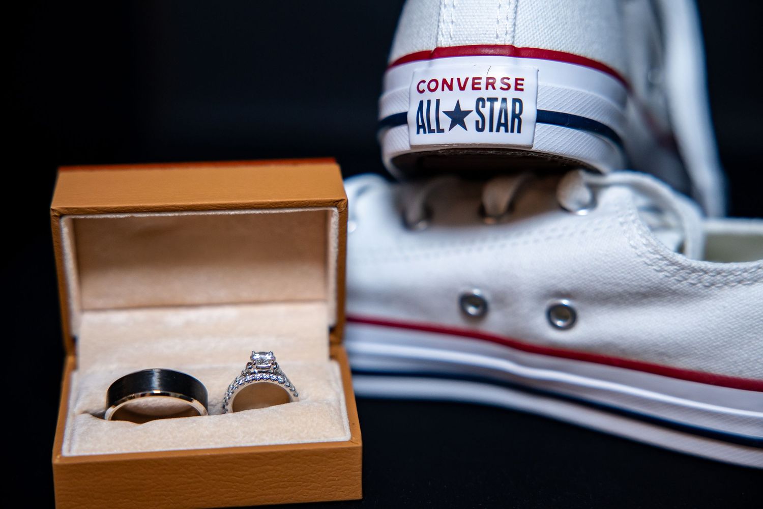 Wedding bands, a black men's ring and a diamond women's ring, in an open tan box beside white Converse All Star sneakers.