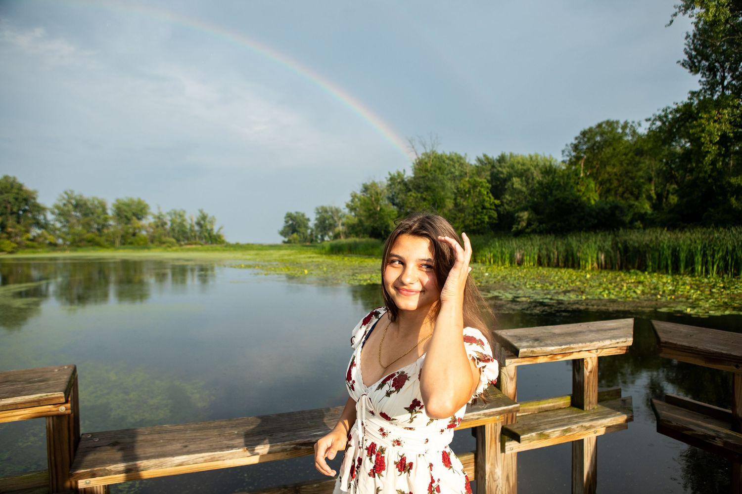 A person in a floral dress enjoys a scenic view from a wooden dock overlooking a tranquil lake on a sunny day.