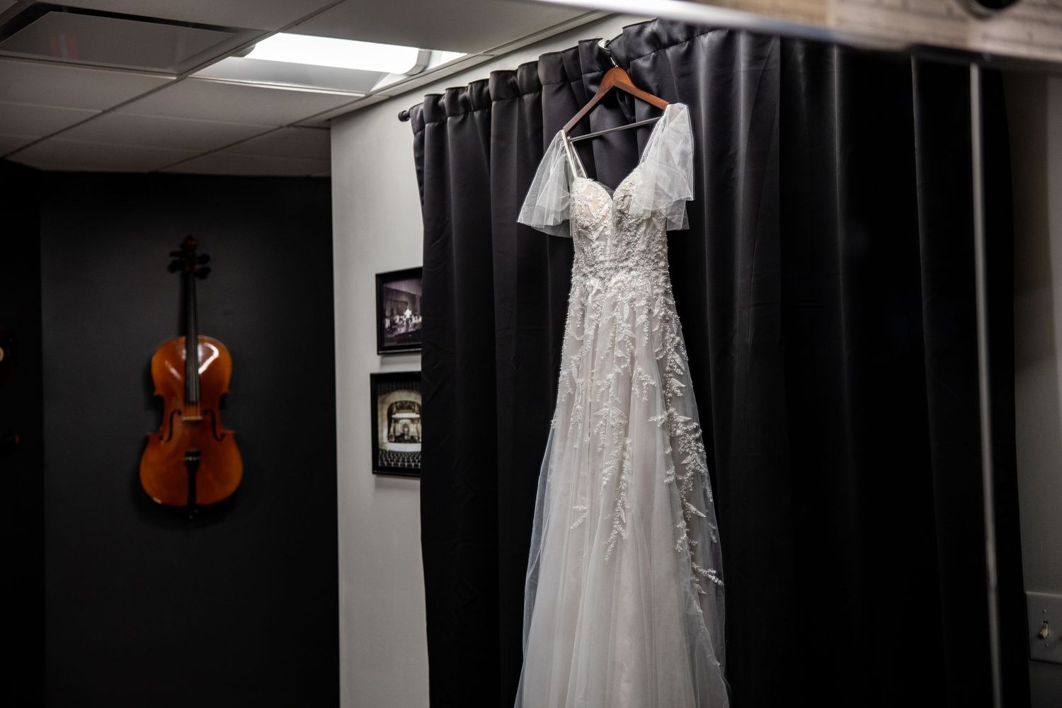 White lace wedding dress hanging on a curtain rod in a room with a violin and framed photos on the wall.
