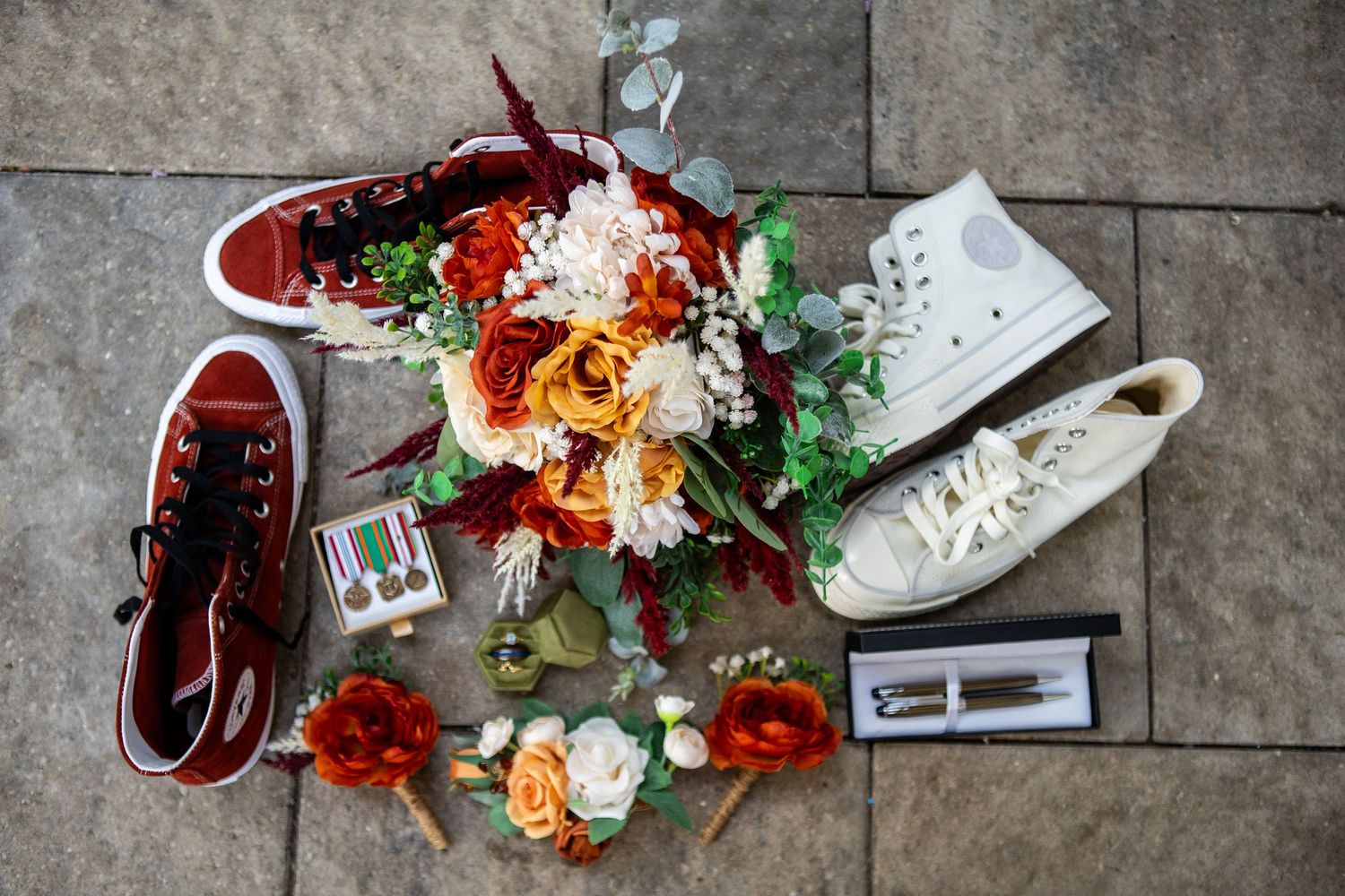 Wedding flat lay with red sneakers, white Converse, floral bouquet, military medals, and flowers on stone floor.