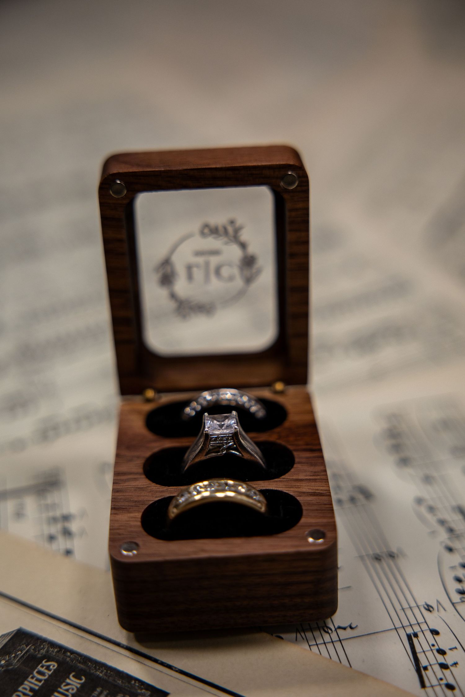 Wooden ring box with diamond engagement ring and wedding band displayed on sheet music.