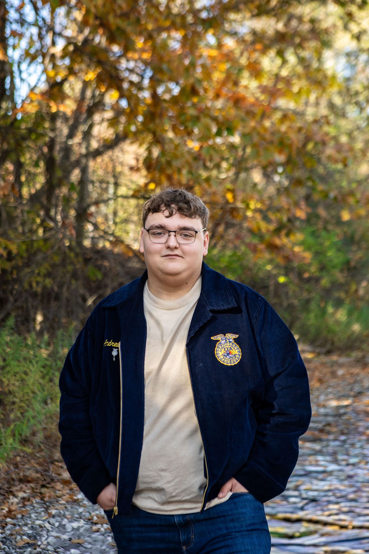 Person wearing navy FFA jacket and light colored shirt stands outdoors among autumn trees.