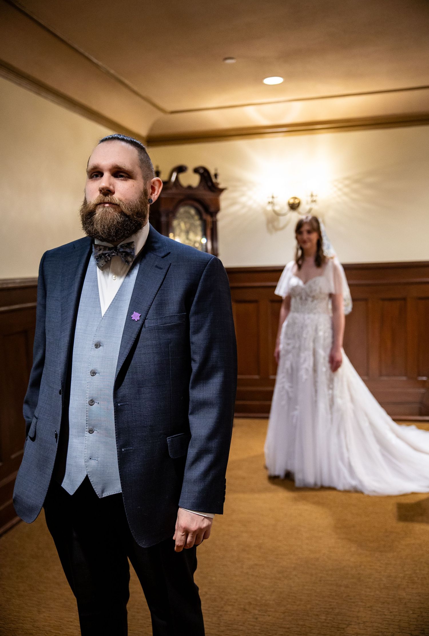 Groom in navy suit with bow tie stands in foreground as bride in white gown waits behind in elegant room.