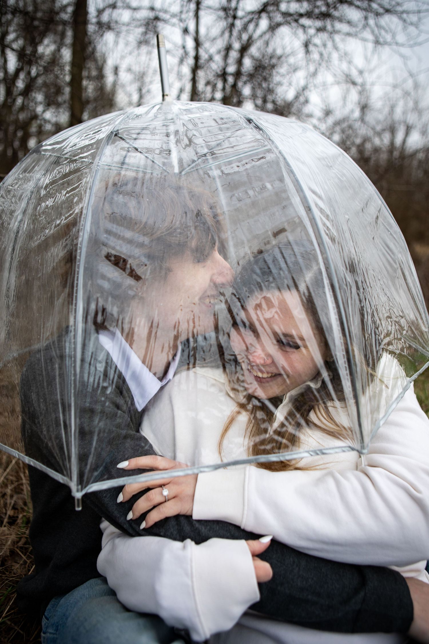 A couple laughs together while sheltering under a transparent umbrella outdoors.