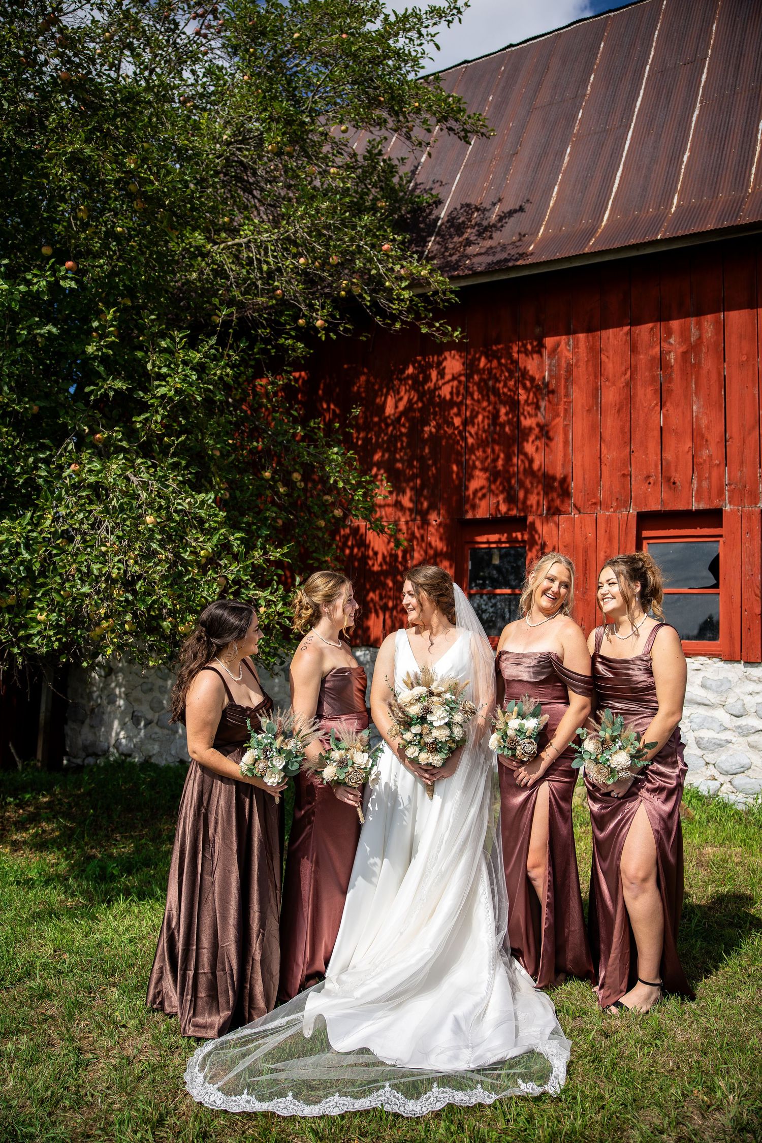 Bride in a long veil holds a bouquet while smiling with four bridesmaids in brown satin dresses beside a red barn.