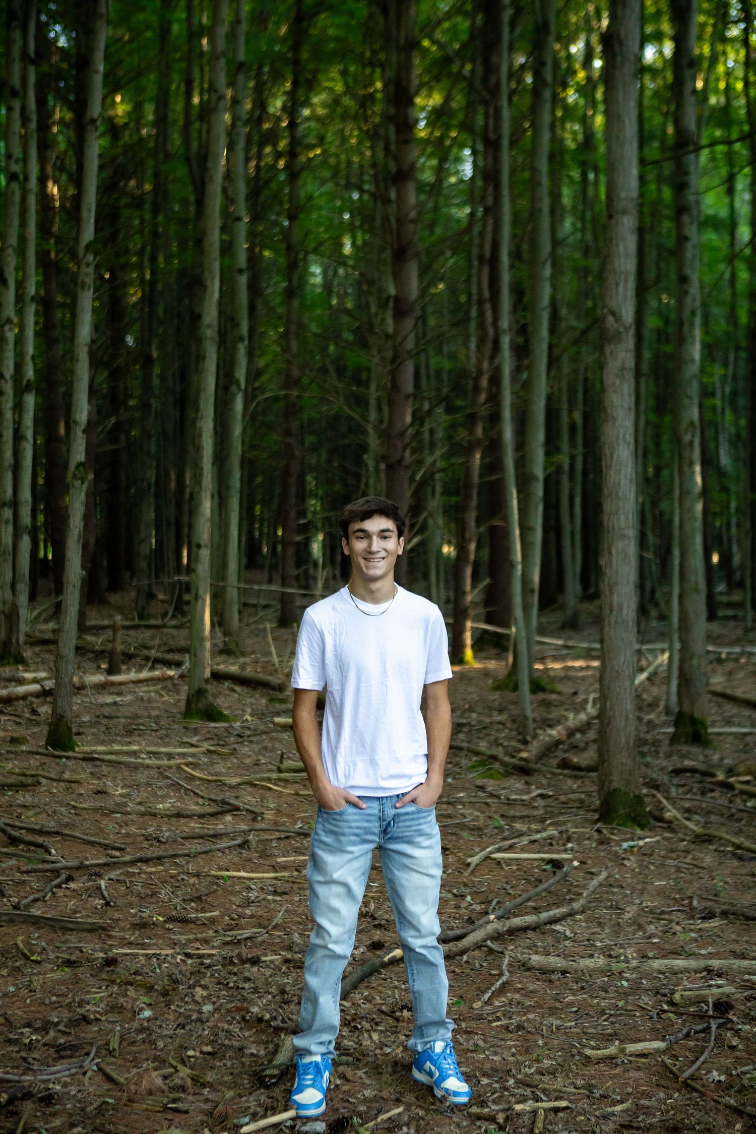 A person wearing blue jeans and a white t-shirt stands in a wooded forest area with tall trees in the background.