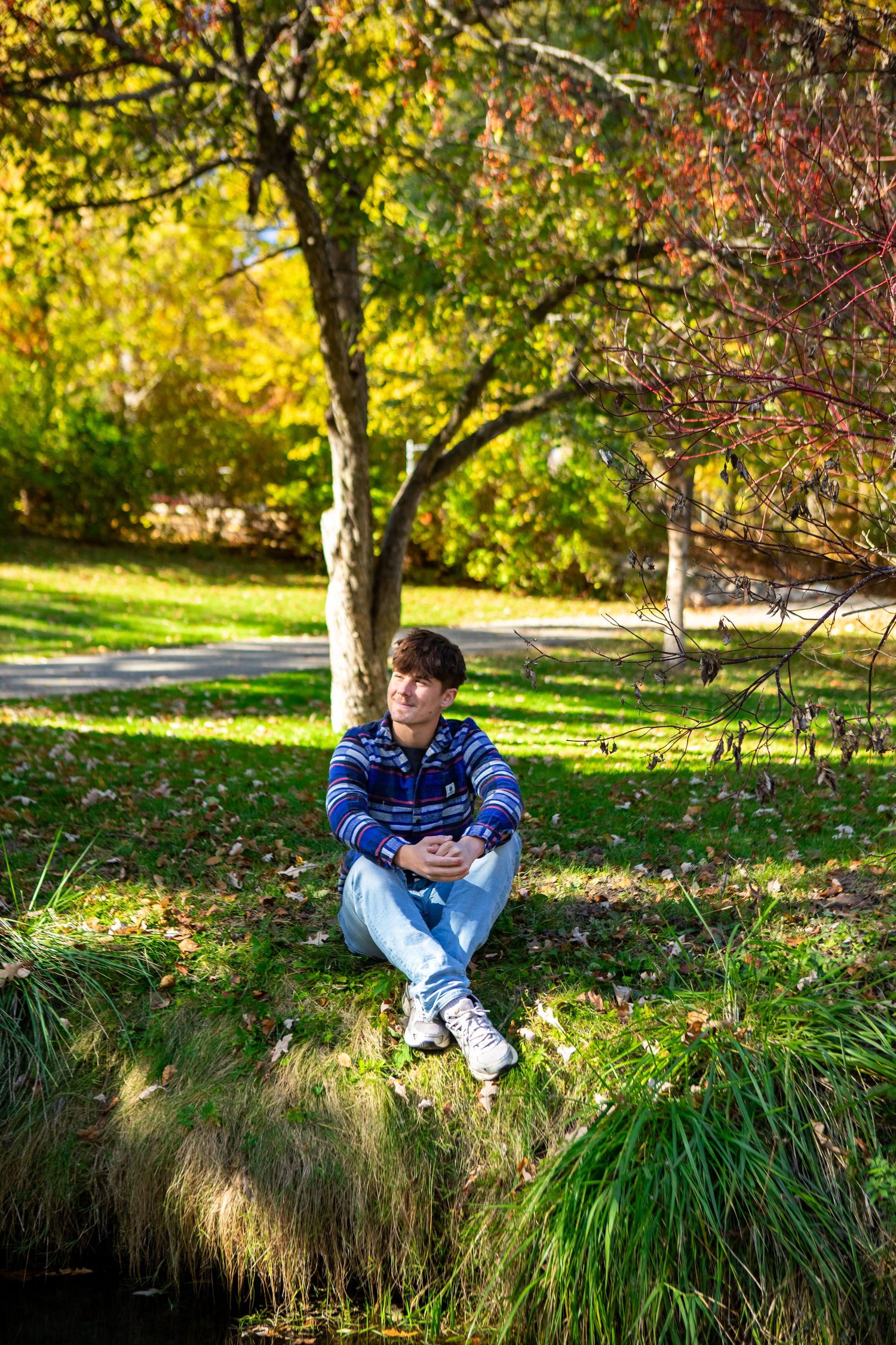 Young man sitting on a grassy bank in a colorful autumn park, wearing jeans and a striped hoodie.