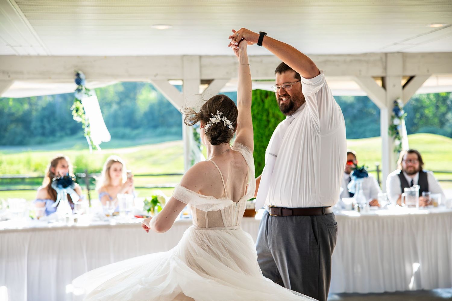 Bride in a white lace gown and groom in a white shirt share a first dance under a pavilion while wedding guests watch from tables.