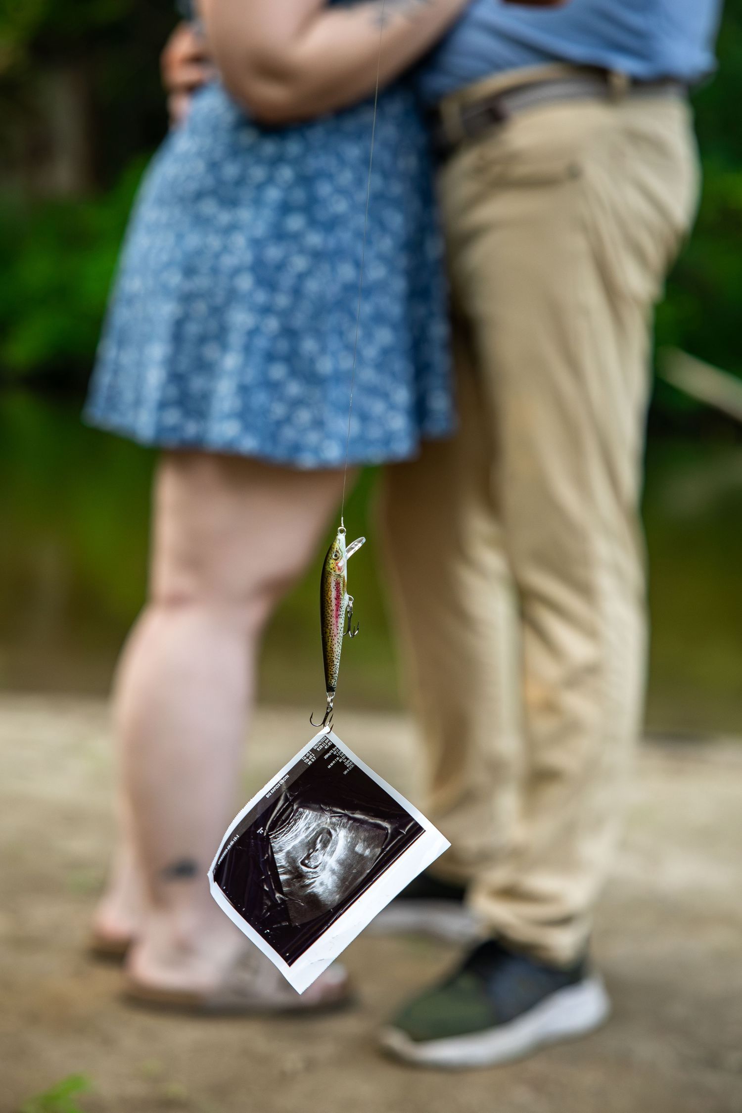 Couple holding fishing lure with ultrasound photo announcing pregnancy outdoors near water.