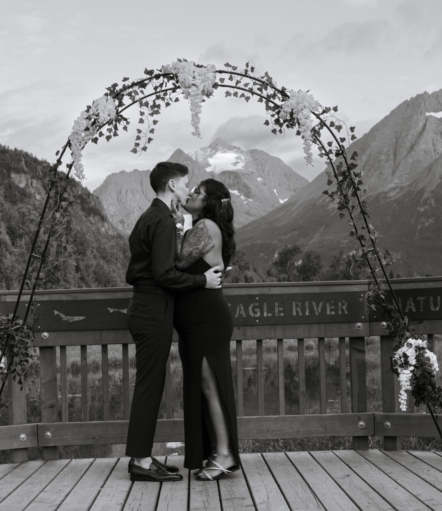 Black and white elopement ceremony photo of a couple kissing beneath a floral arch with Alaska mountains.