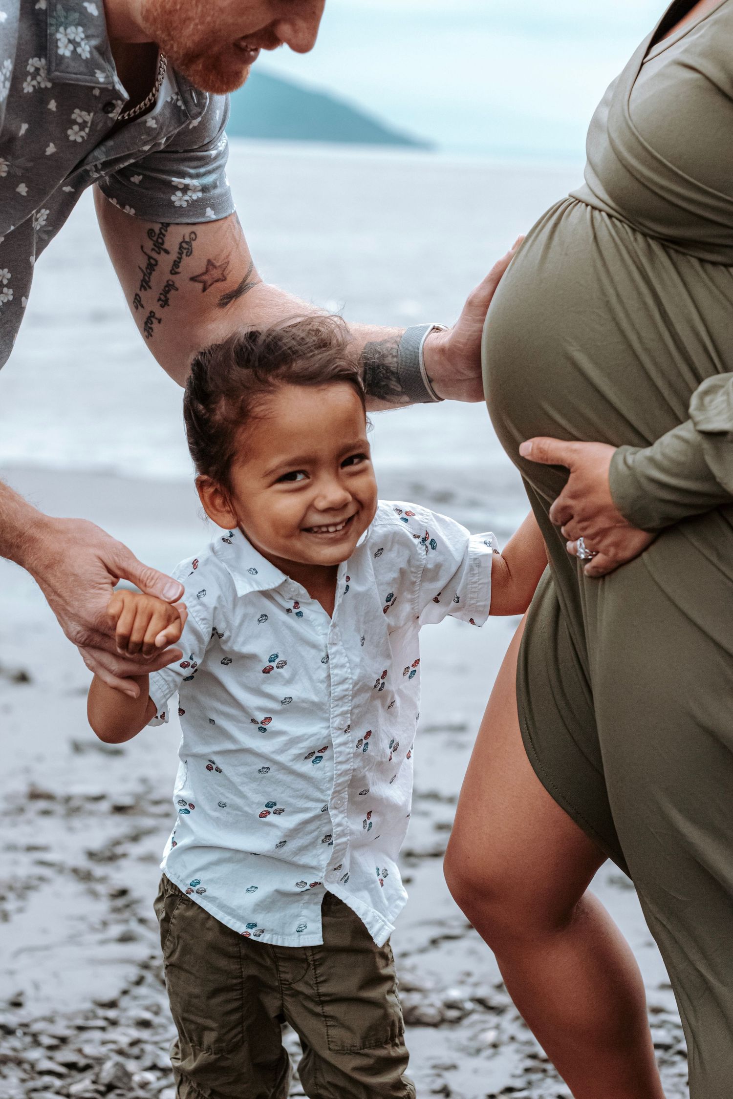 Smiling toddler holding hands with parents during a candid family photo session by the water.