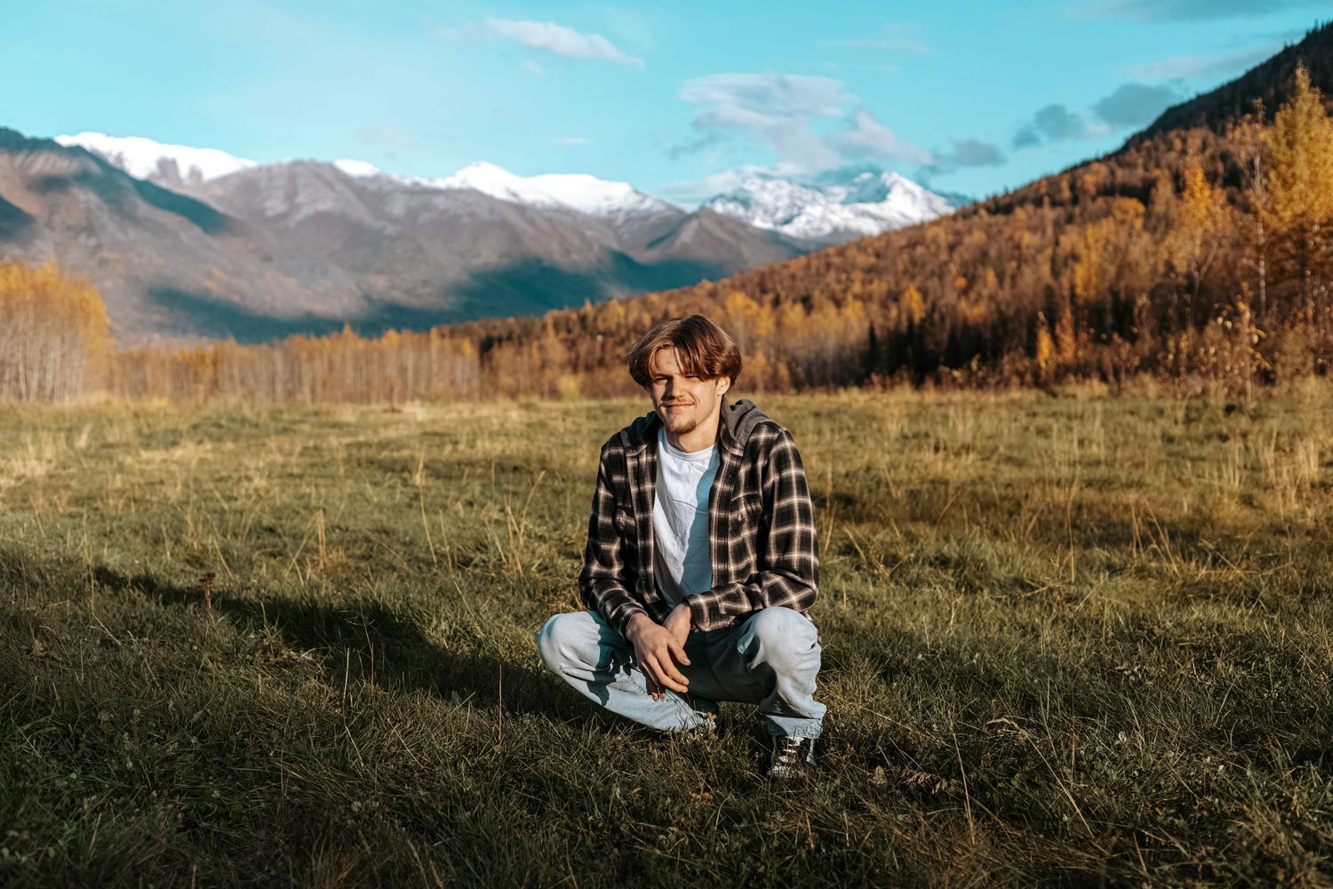 Senior portrait of a young man sitting in an open field with Alaskan mountain scenery behind him.