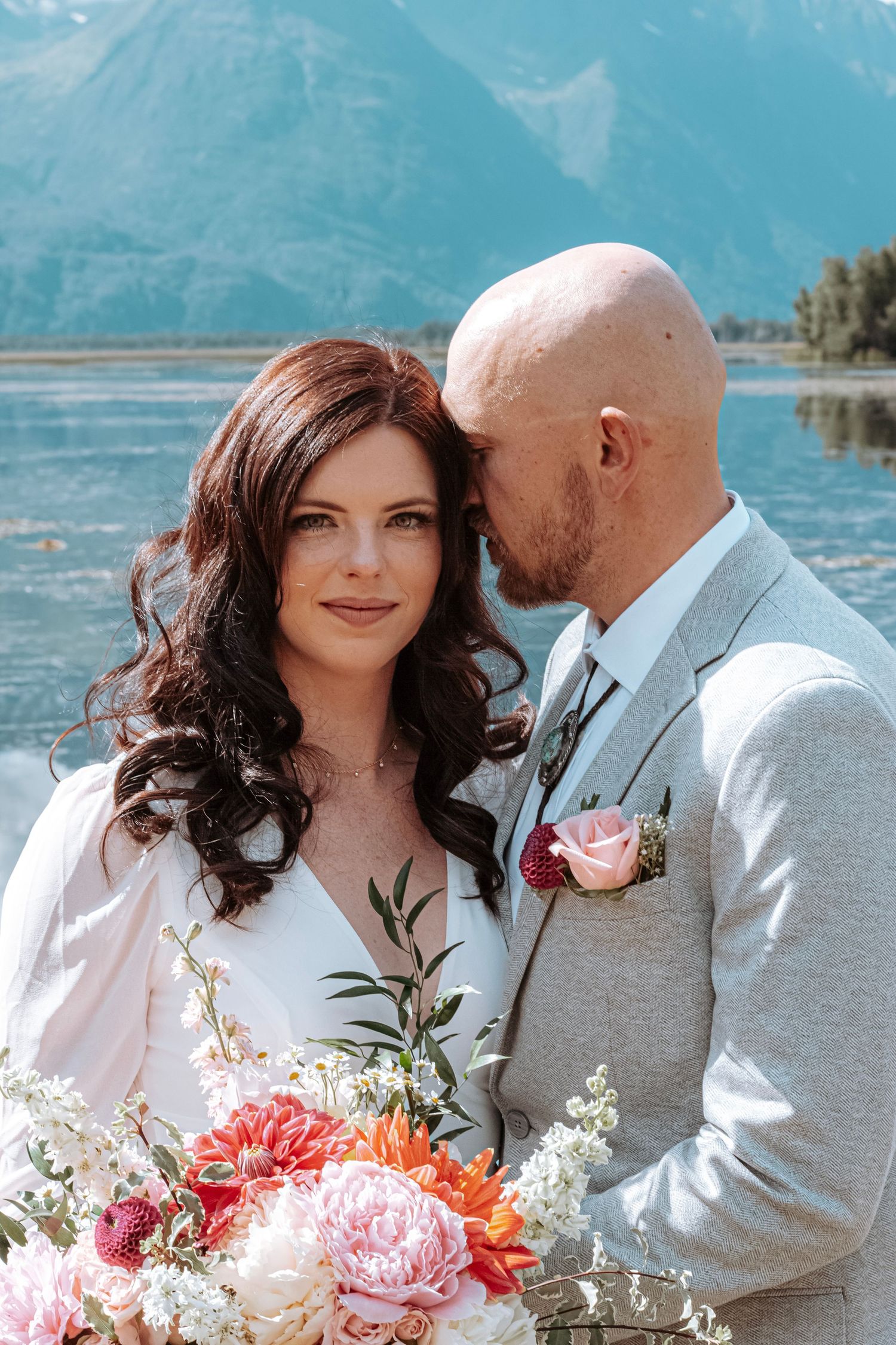 Bride and groom standing together by the water during an intimate Alaska wedding, holding a colorful floral bouquet.