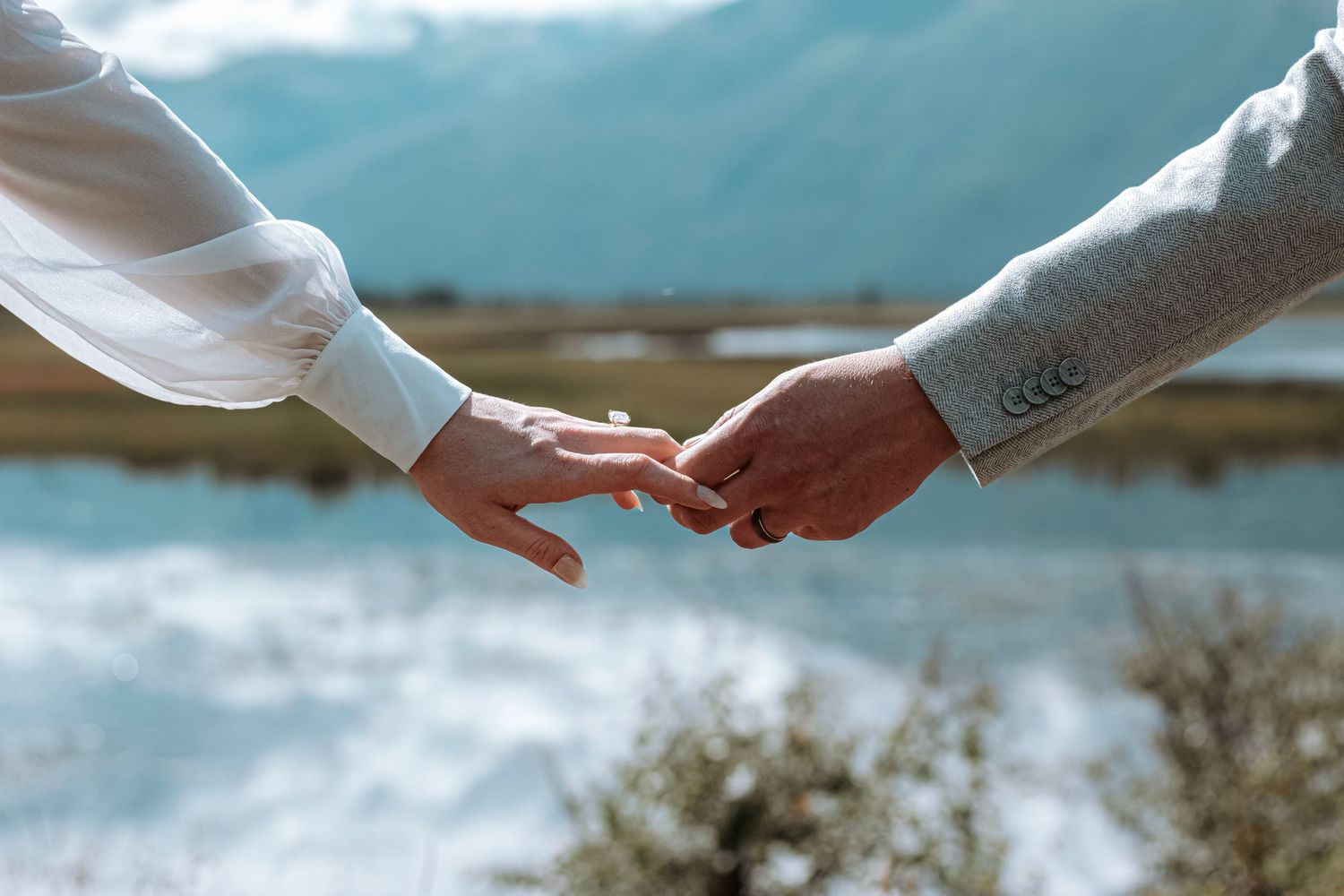 close-up of a couple holding hands with wedding rings, set against a scenic mountain and lake backdrop in Alaska – romantic outdoor wedding