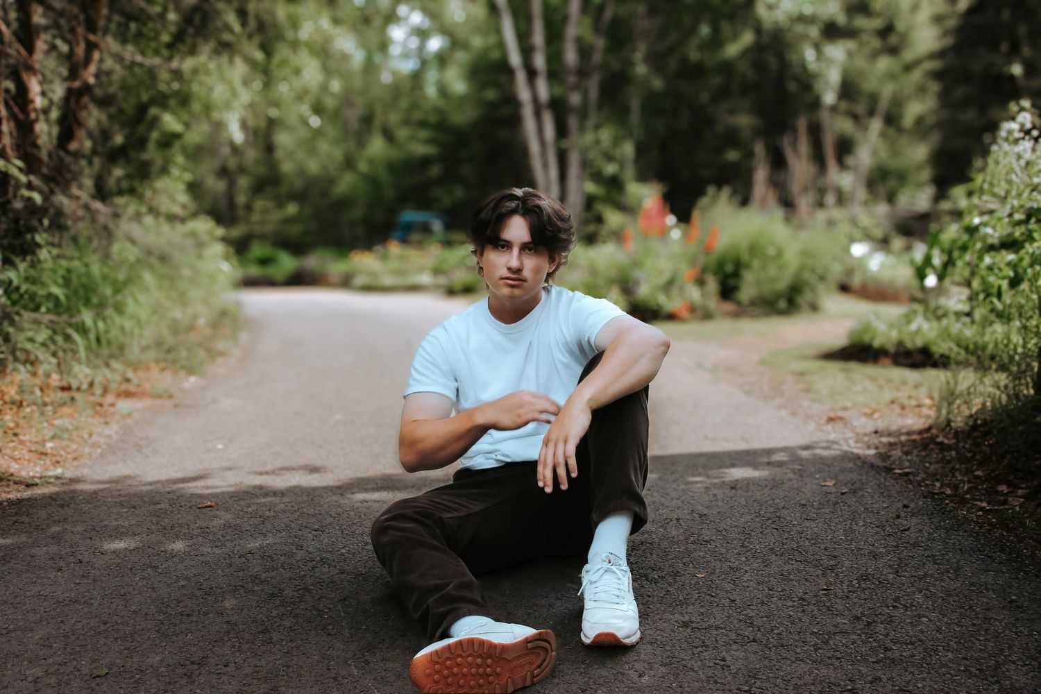 Senior portrait of a young man sitting on a paved path surrounded by greenery during an outdoor senior session in Alaska.