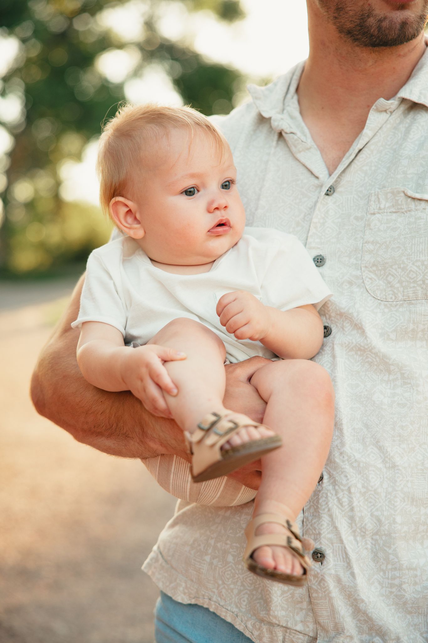 A father holds his adorable baby in a white onesie outdoors in warm golden sunlight.