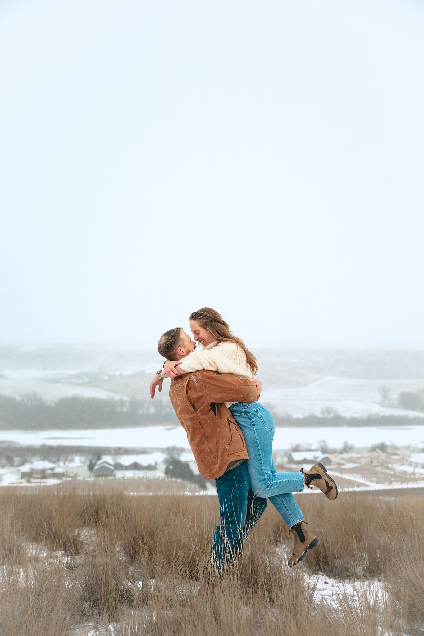 Couple embracing in a snowy winter field, man lifting woman in blue jeans and cream sweater outdoors.