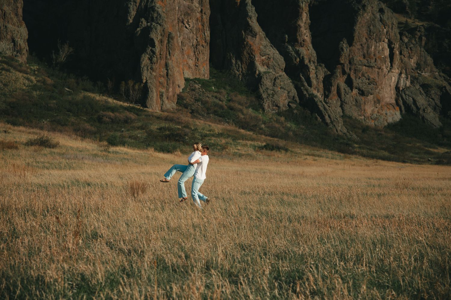 Couple dancing joyfully in a golden meadow with dramatic rocky cliffs in the background.