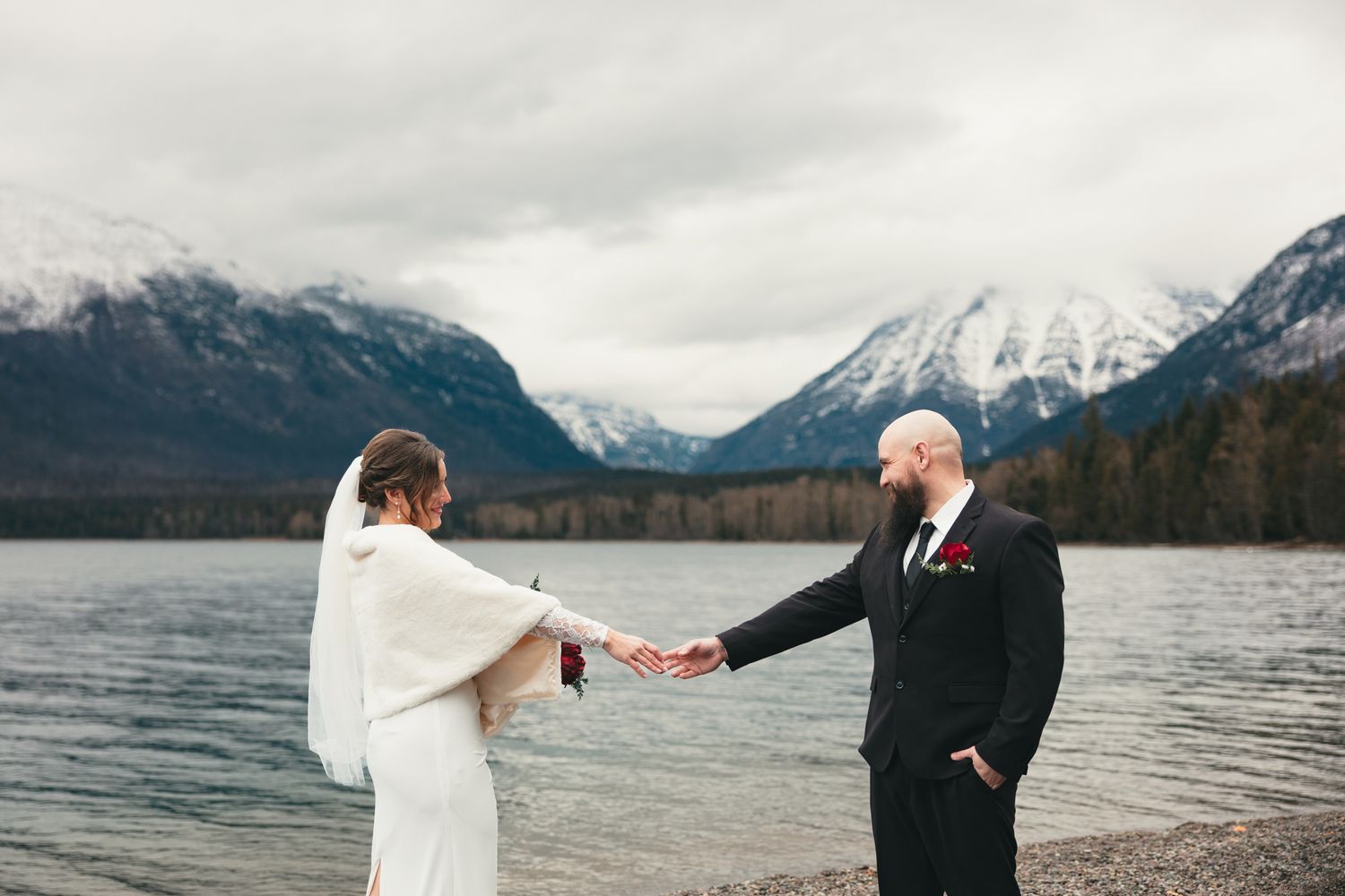 Bride and groom hold hands by a mountain lake with snow-capped peaks, bride in white wrap, groom in black suit.