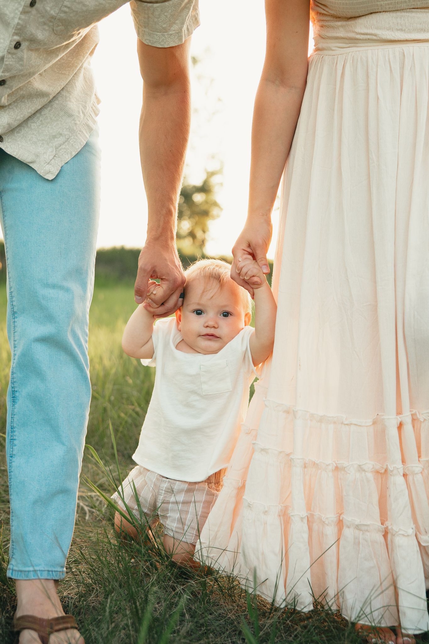 Baby taking first steps outdoors, held by parents hands in sunny field, wearing white onesie.