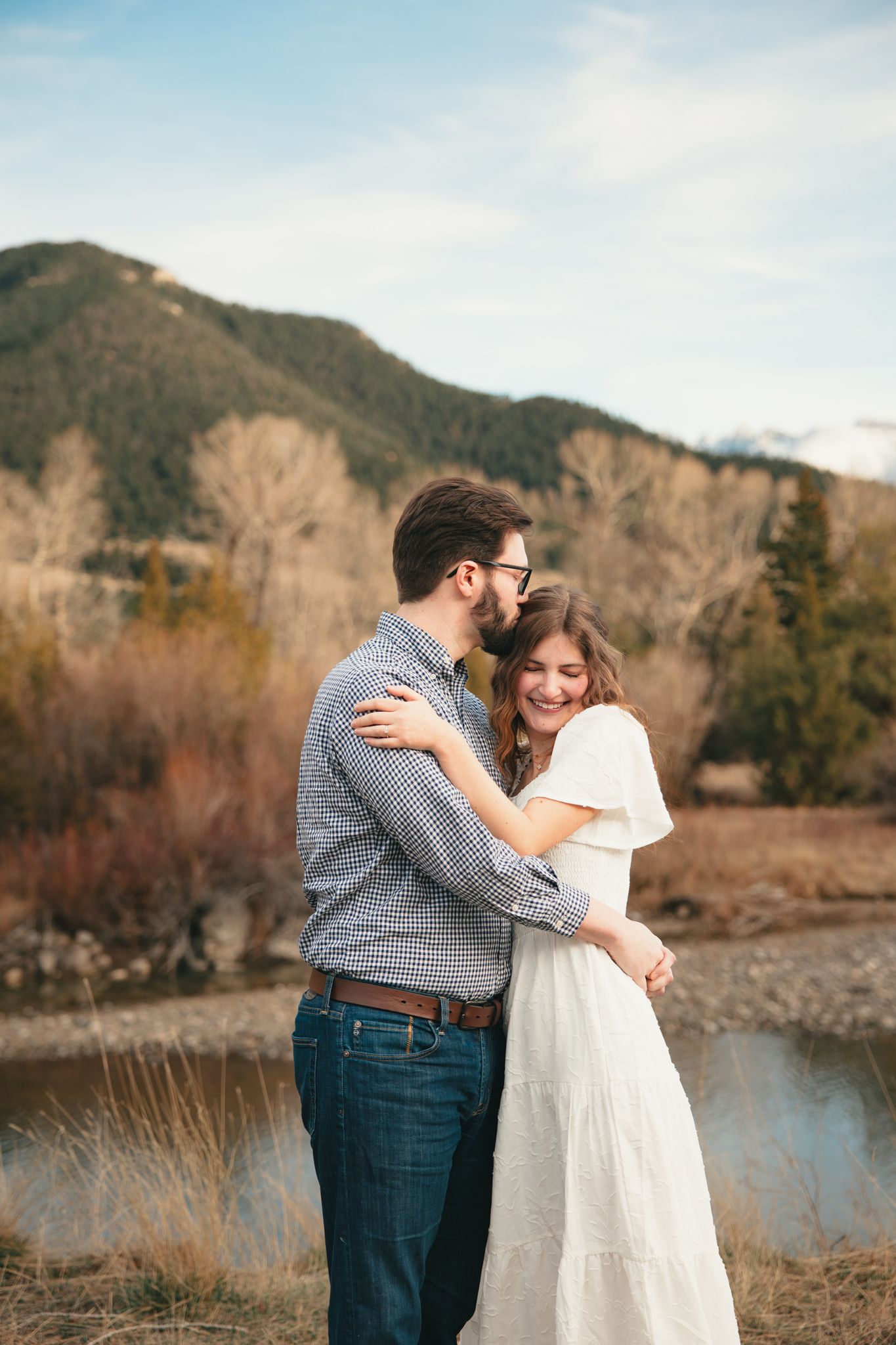 A couple embraces and laughs together outdoors with mountain scenery and a river in the background.