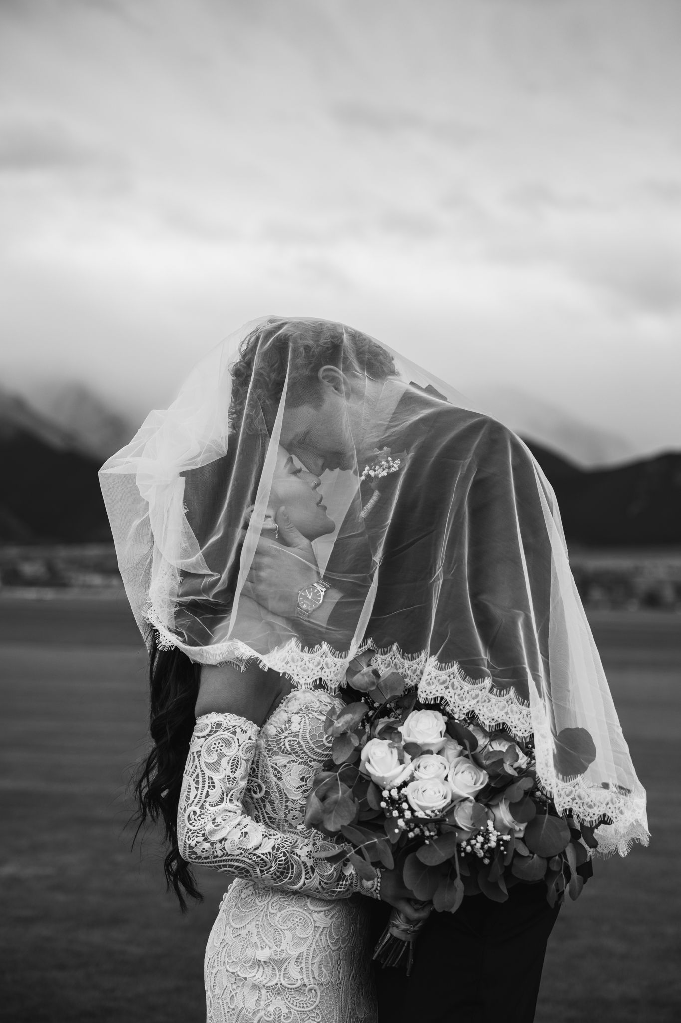 Black and white photo of bride and groom kissing under veil, holding bouquet, with mountain backdrop.