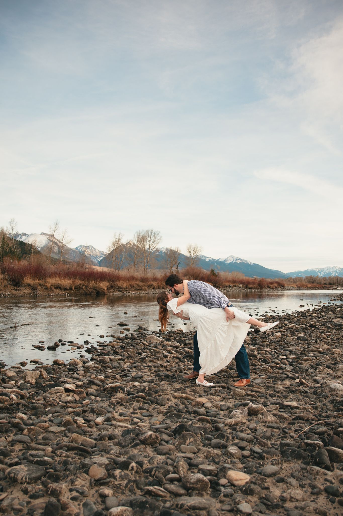 Couple sharing romantic dip kiss on rocky riverbank with snow-capped mountains and bare trees in background.