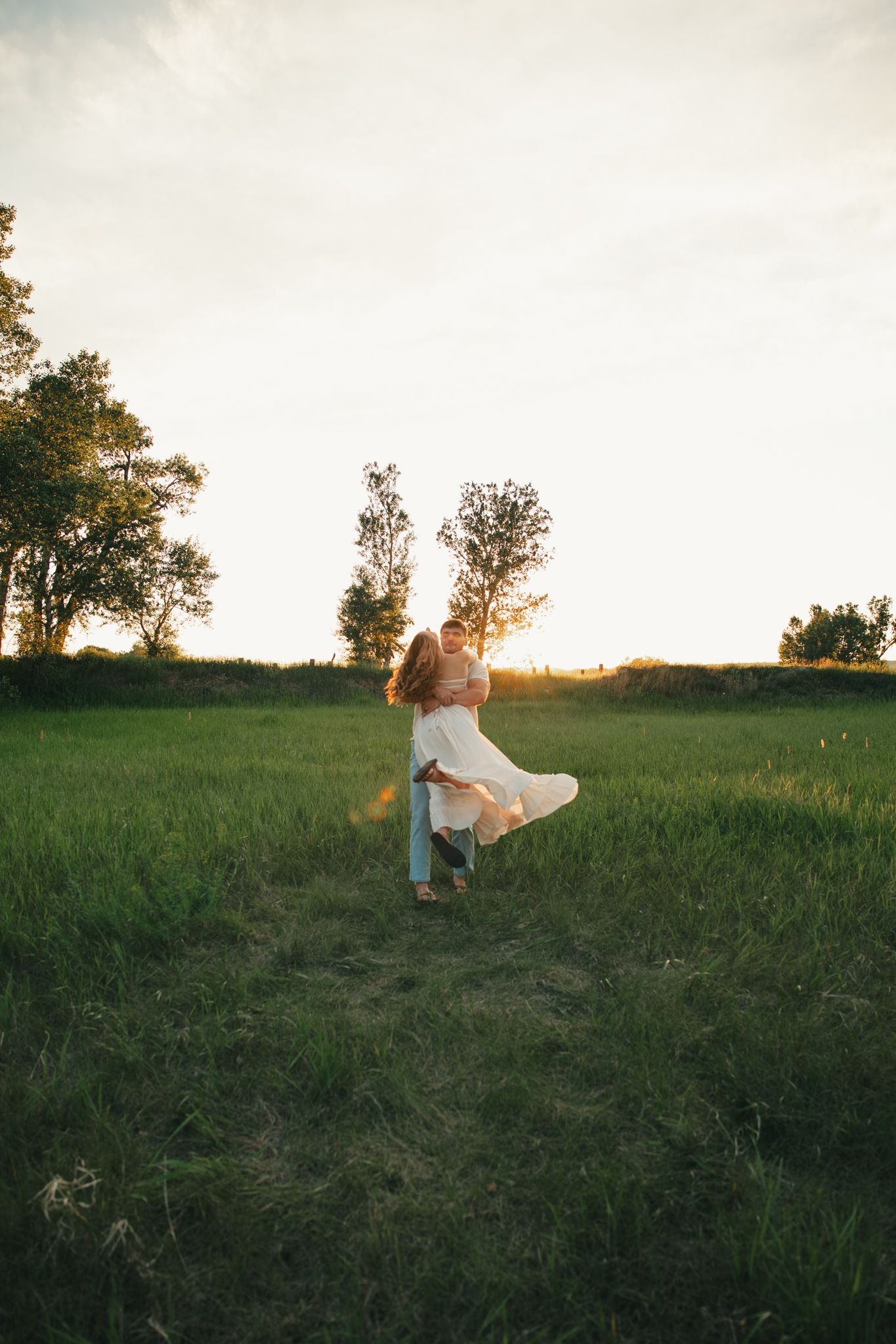 Couple embracing in a lush green field at golden sunset, woman in white dress being lifted by partner.