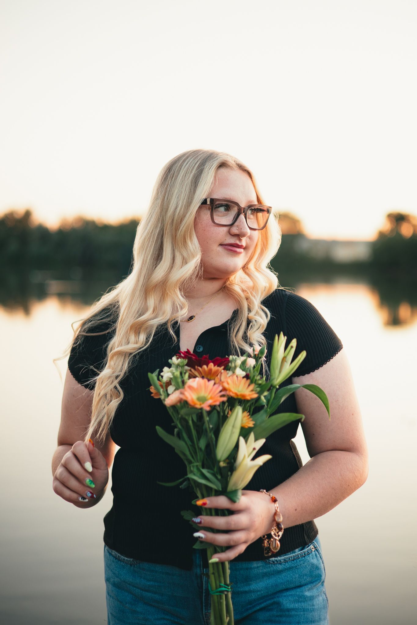 Senior in black top and jeans holds colorful bouquet of lilies and sunflowers by lakeside at sunset. photo, Great Falls, MT