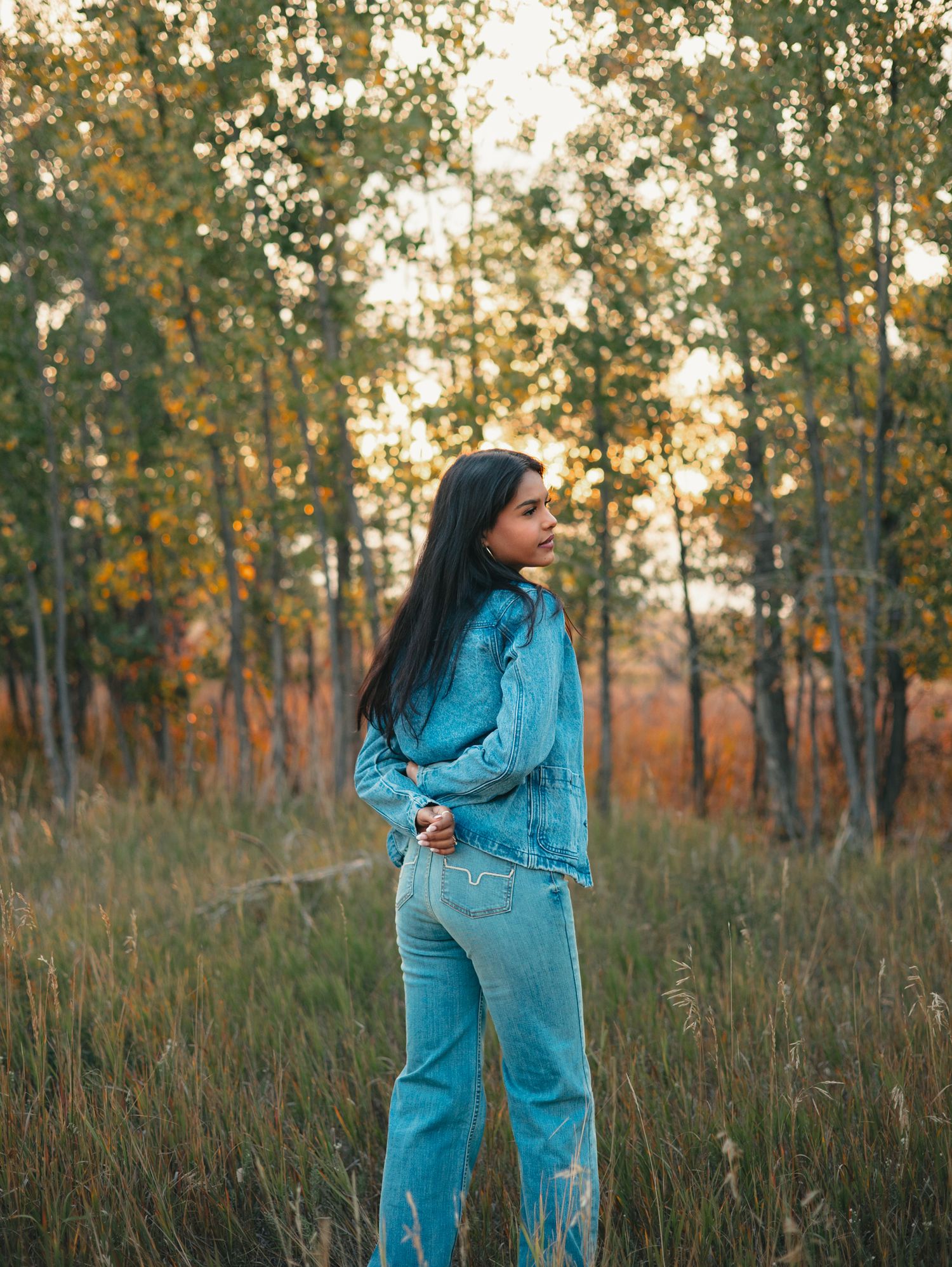 A senior in a bright turquoise jumpsuit poses gracefully in a sunlit autumn field with tall grass and trees. photo, Great Falls, MT